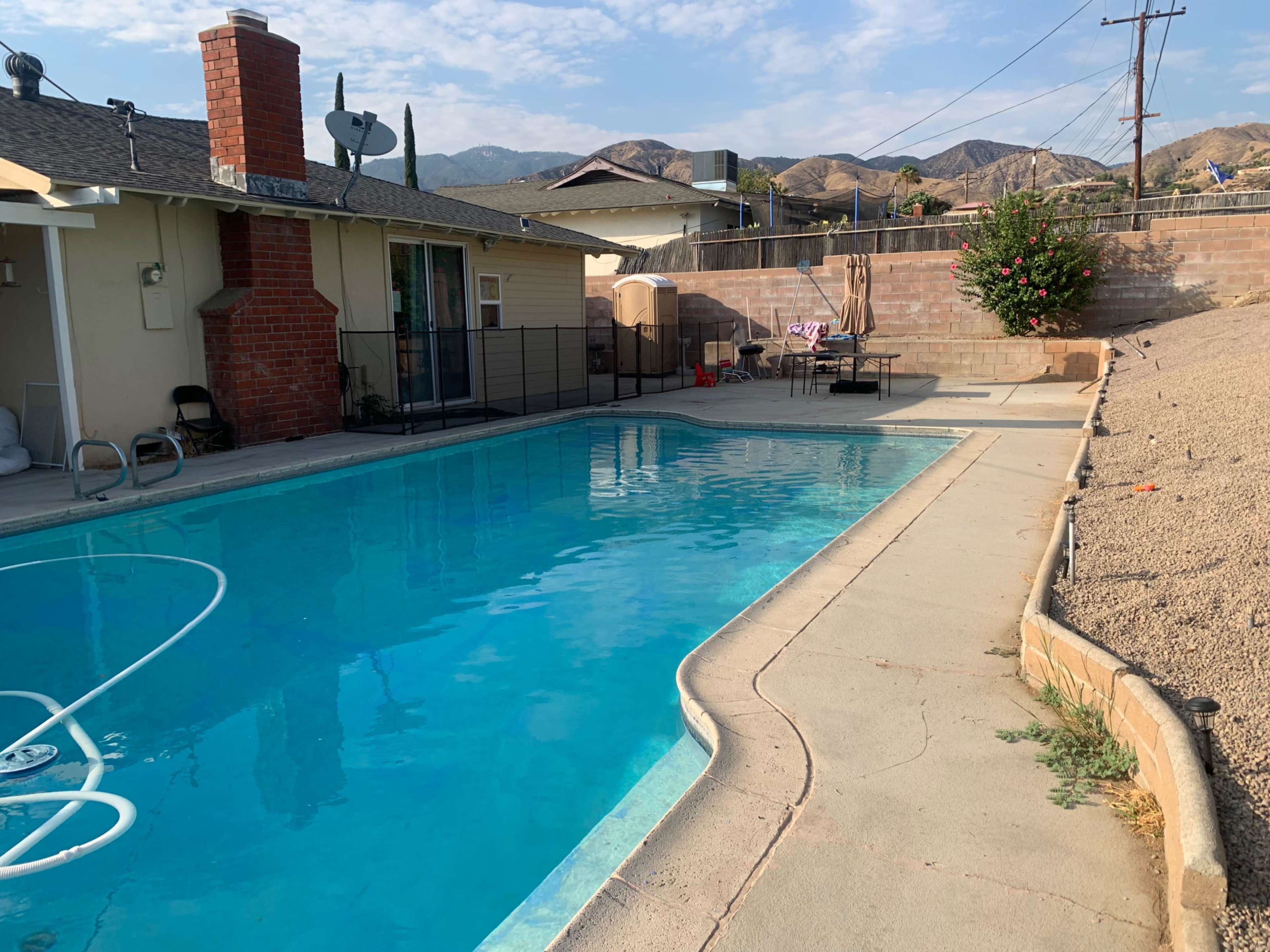 A swimming pool surrounded by a concrete deck and a fence, with a house and hills visible in the background.