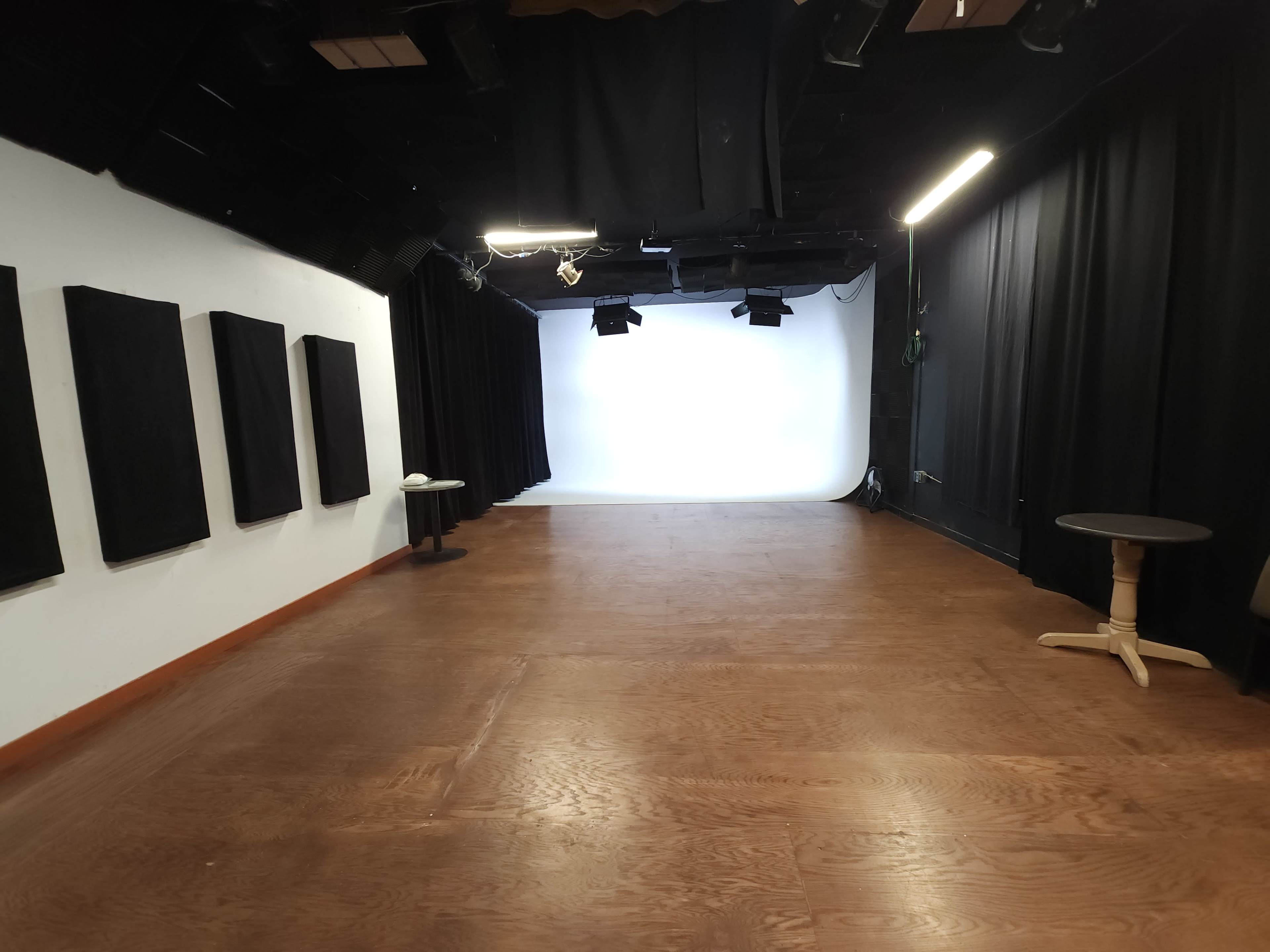 An empty studio space with a white backdrop and black curtains, featuring wooden flooring and two small tables.