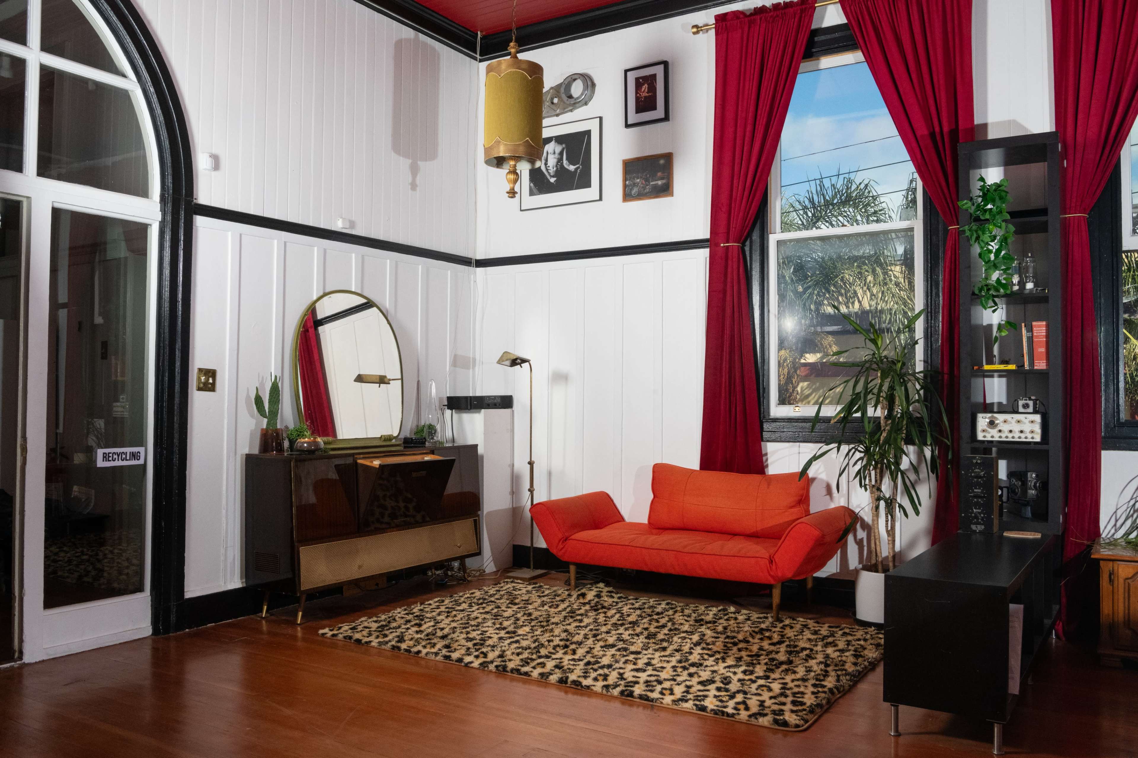 A stylish living room featuring an orange sofa, a large mirror, and decorative plants, with white walls and red curtains.