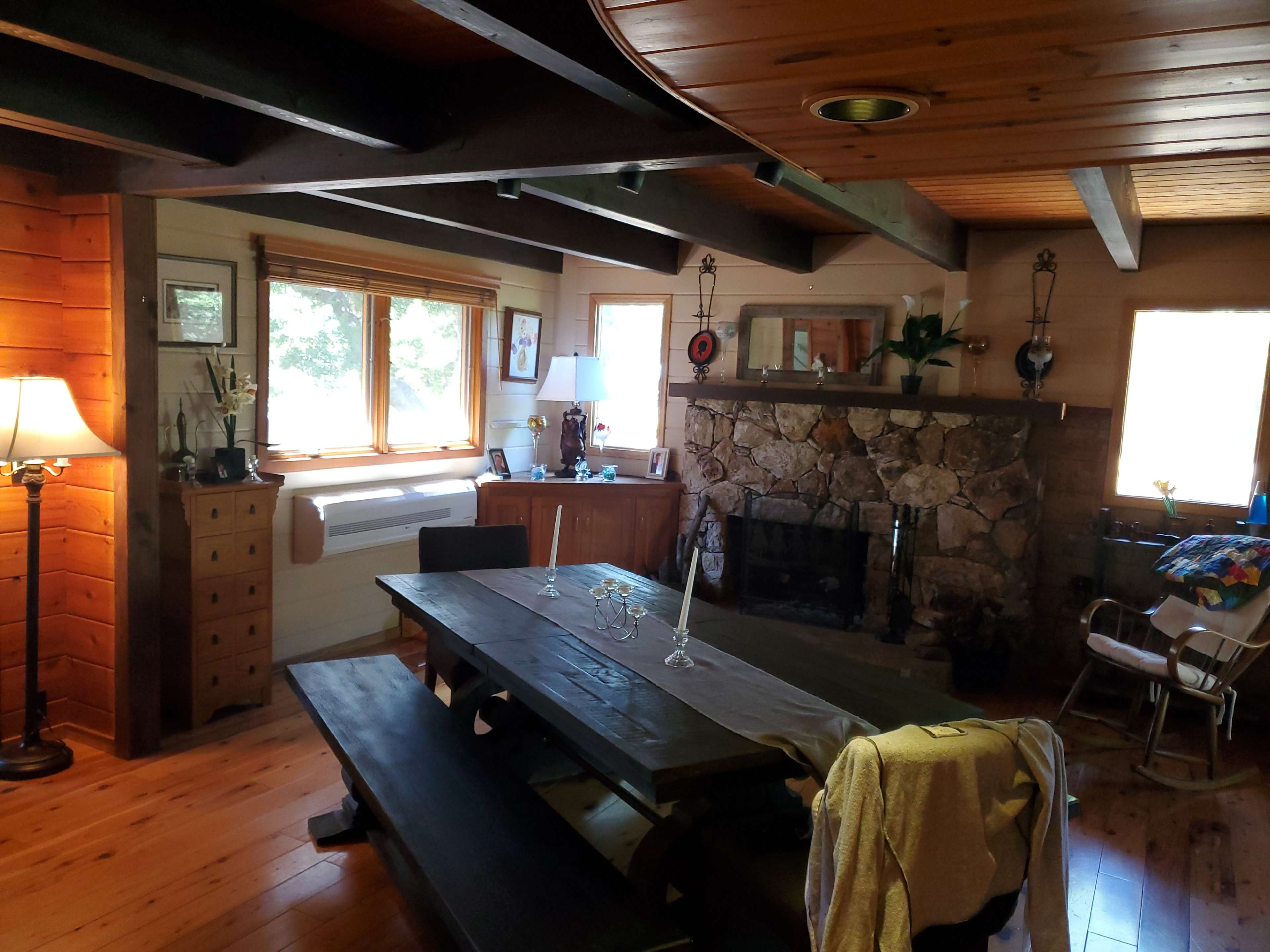 A wooden dining area with a large table and stone fireplace, illuminated by natural light from the windows.