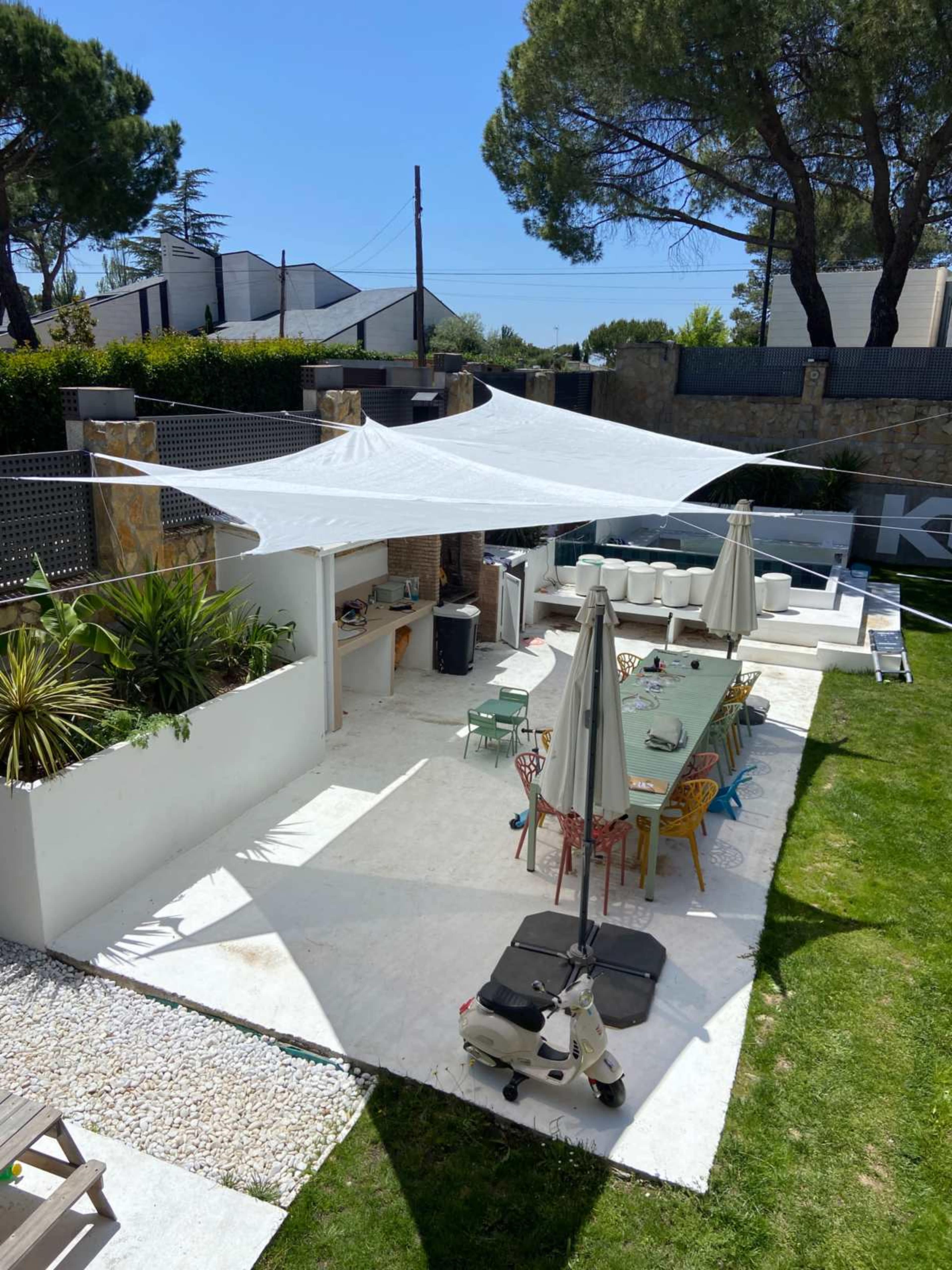 The image shows a bright outdoor dining area with a large white shade sail overhead, surrounded by greenery and colorful chairs.