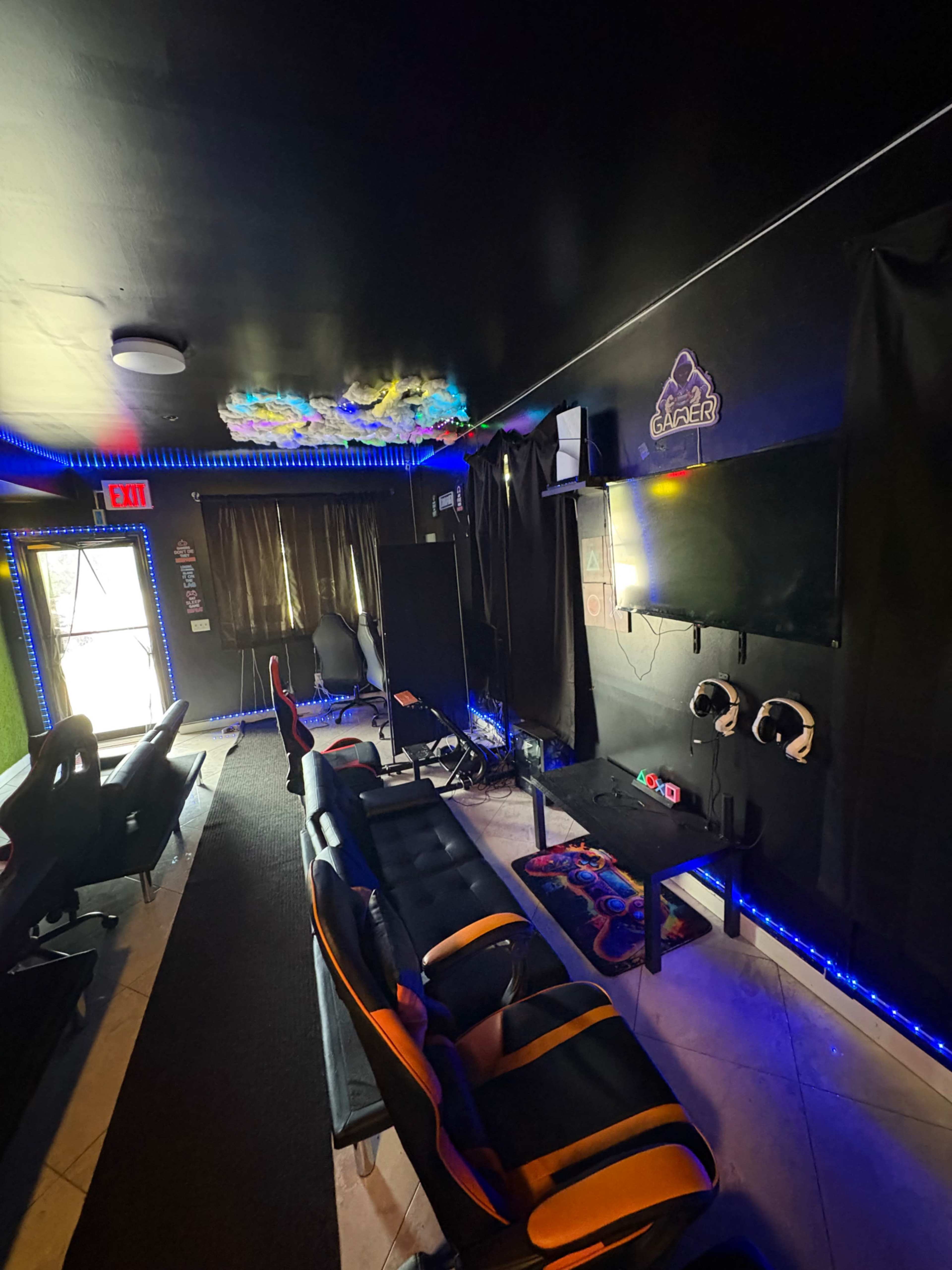 The image shows a gaming lounge with black walls, colorful LED lights, gaming chairs, and a large wall-mounted TV.