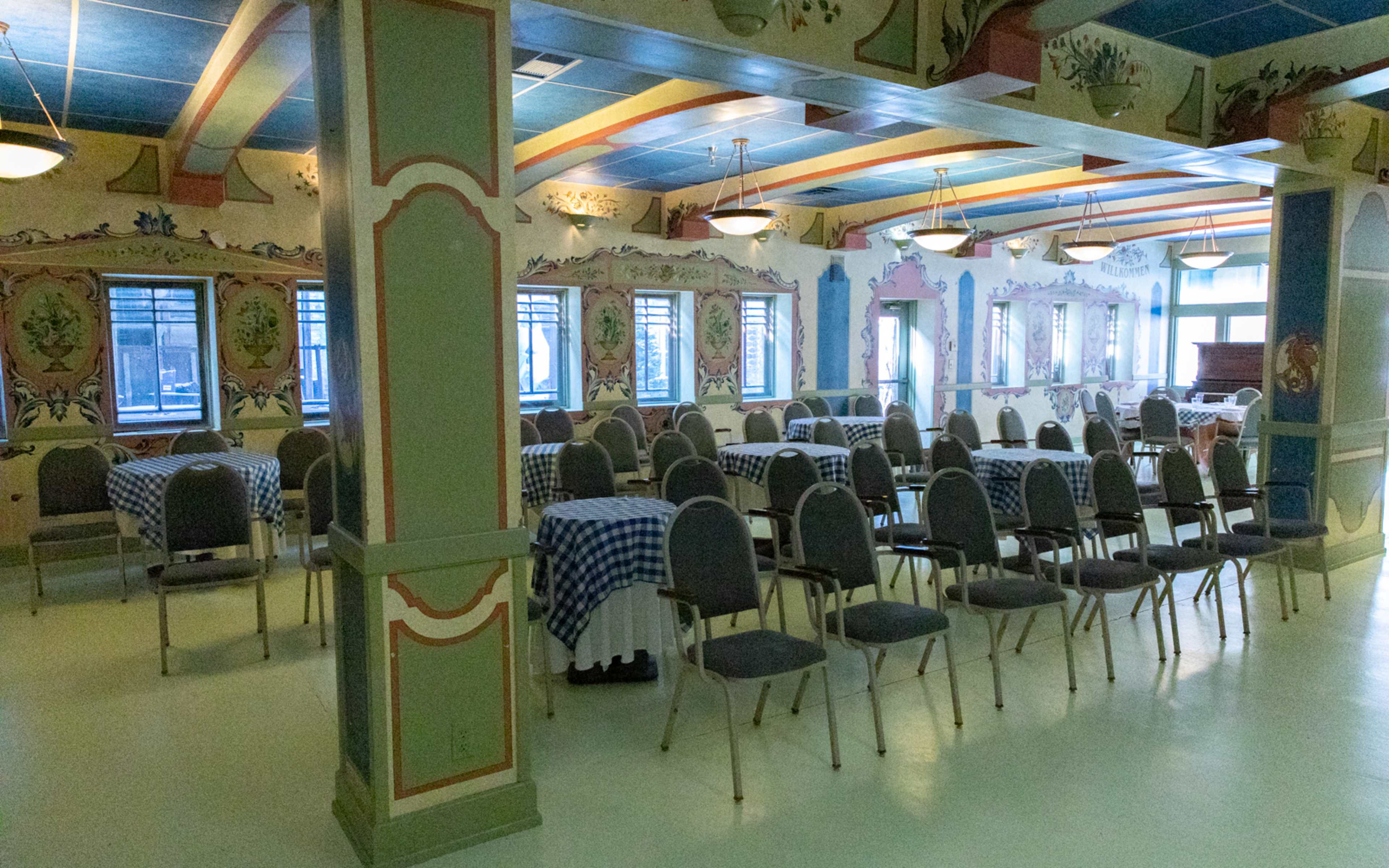 An empty dining room with tables covered in blue checkered cloths and rows of chairs arranged around them, surrounded by colorful murals on the walls.