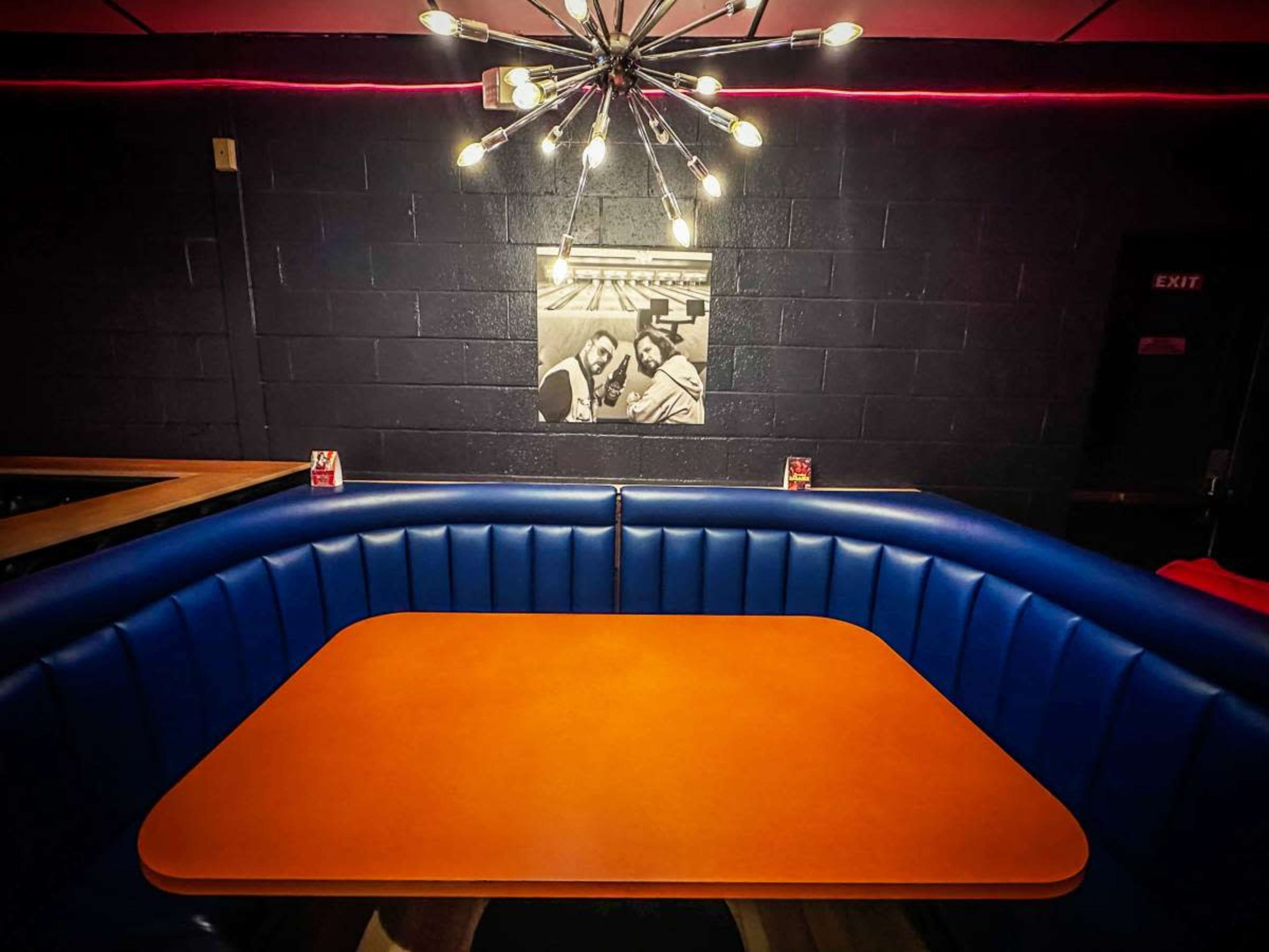 The image shows a retro-style booth with blue upholstery and an orange table, illuminated by a starburst light fixture, against a black wall featuring a framed black-and-white photograph.
