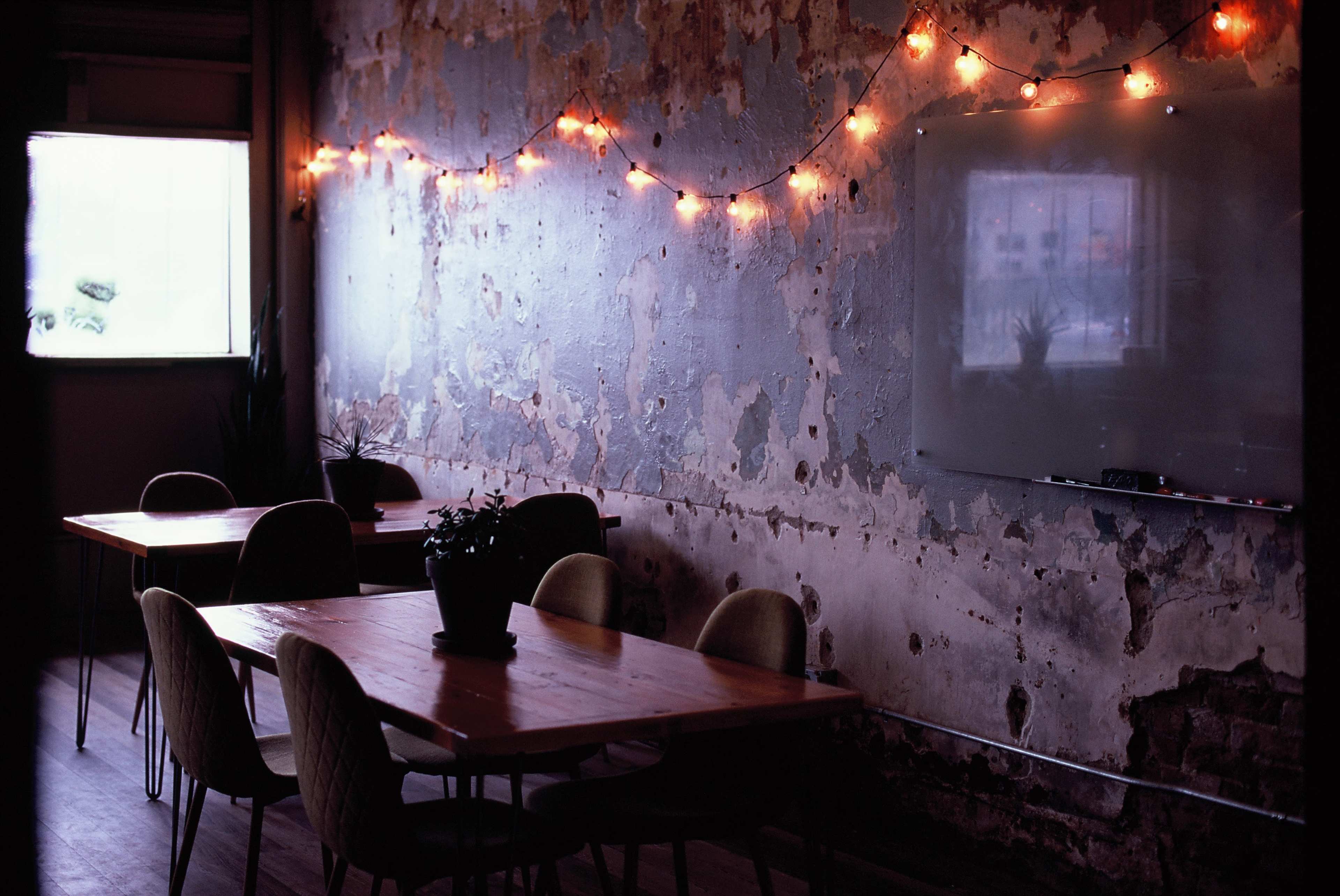 A dimly lit room with peeling walls, wooden tables, and a whiteboard, adorned with string lights along the ceiling.