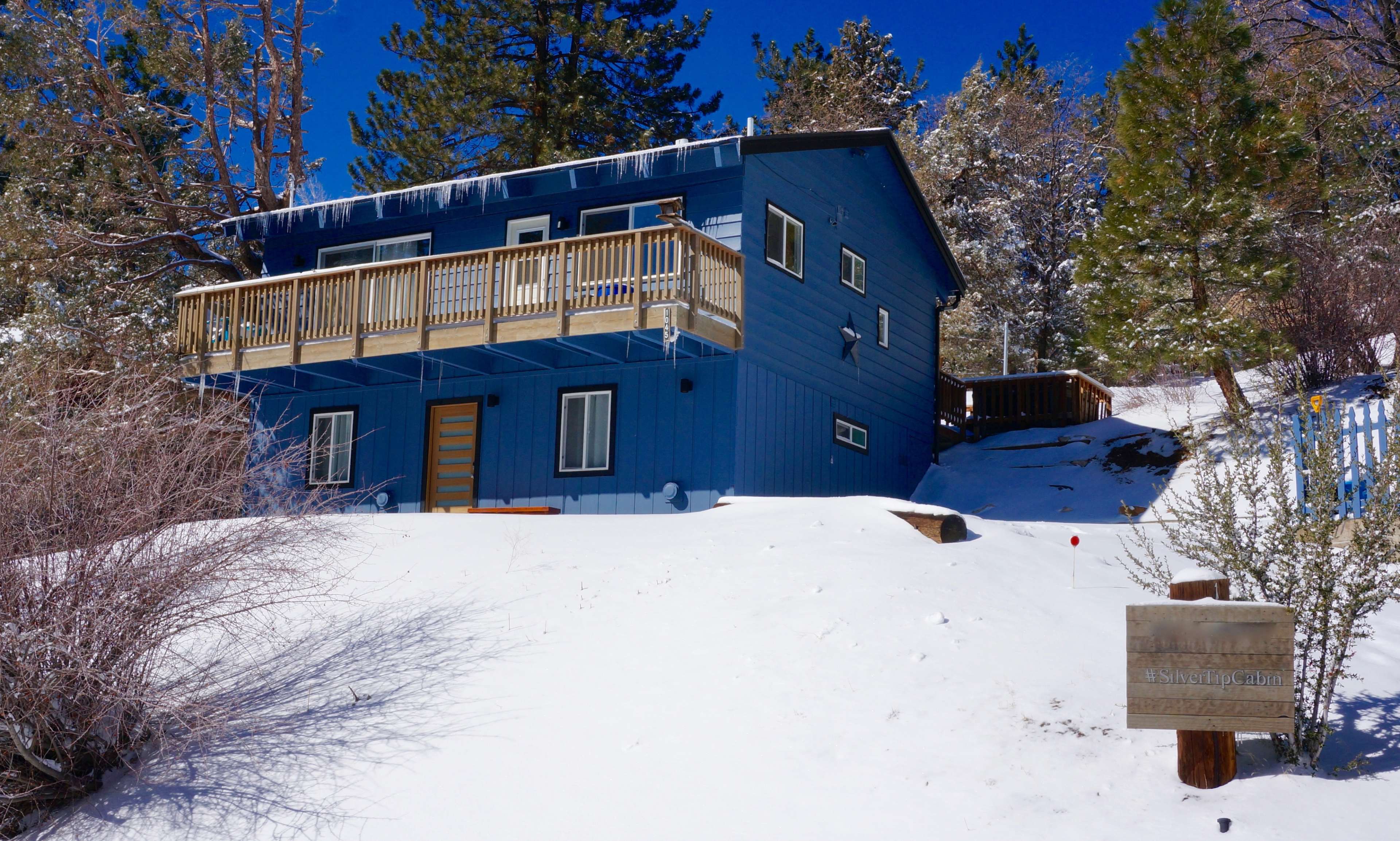 A blue two-story house sits on a snowy hillside, surrounded by trees and a sign at the front.