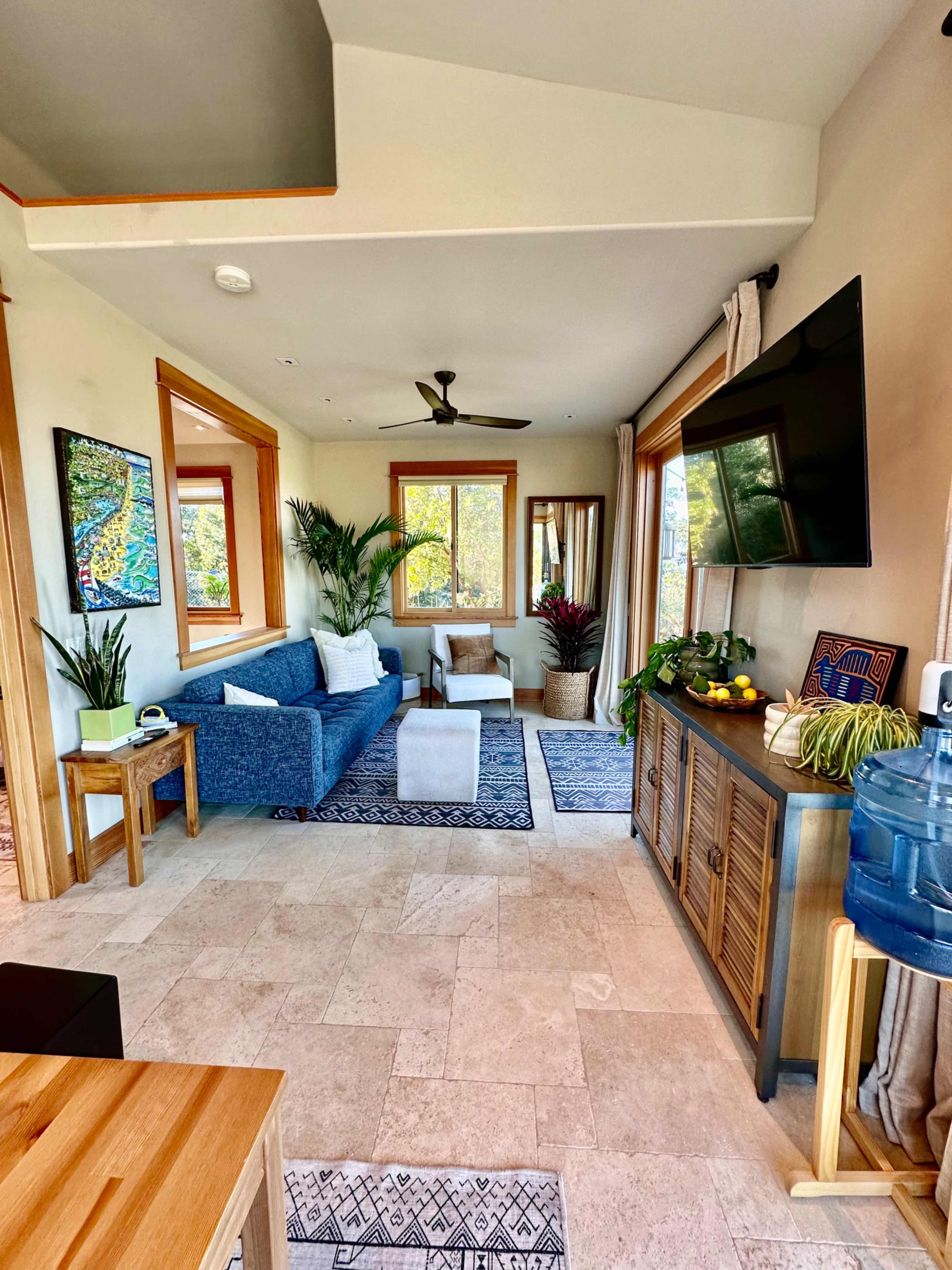 The image shows a bright living room with a blue sofa, a white armchair, large windows, and potted plants.