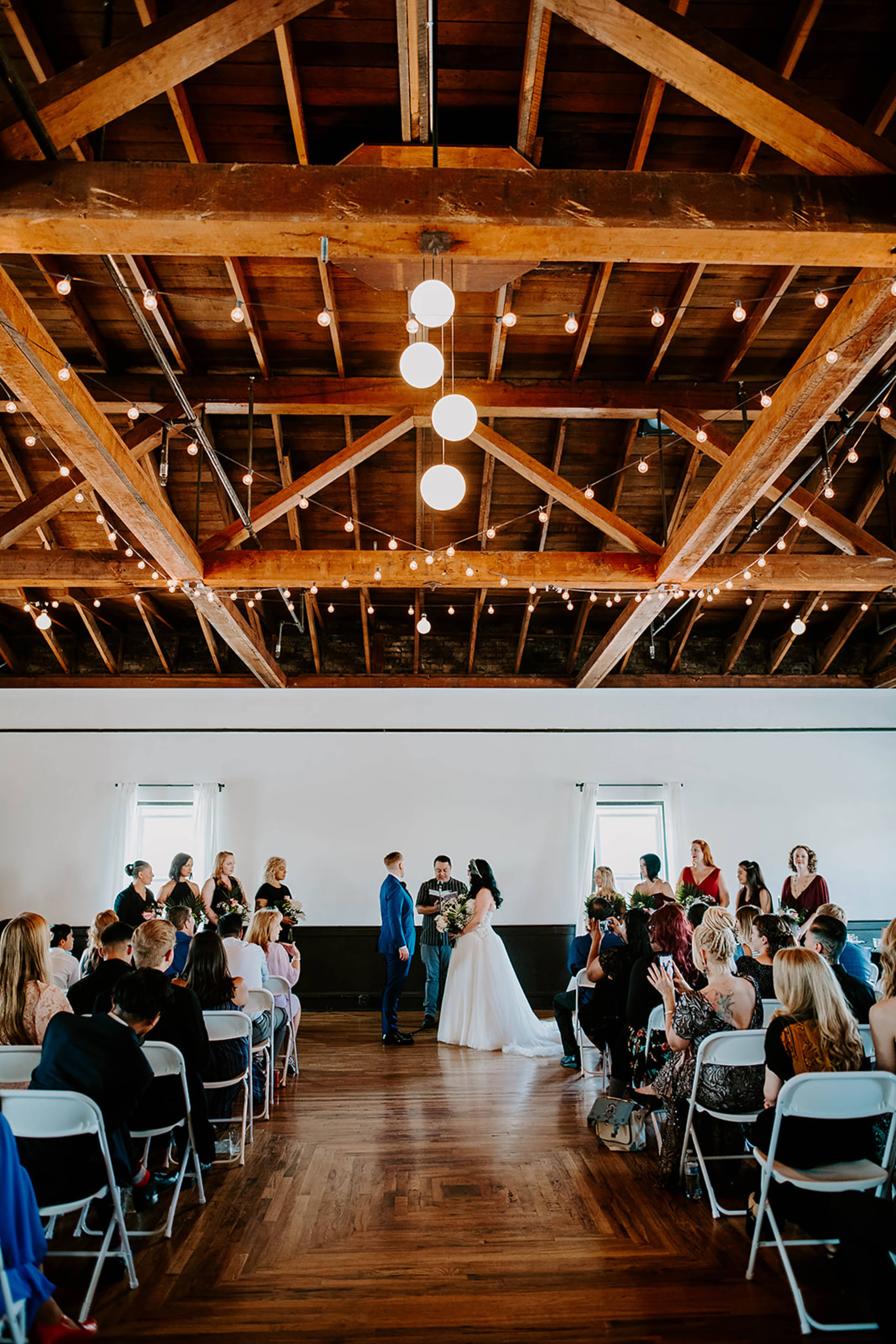 A wedding ceremony takes place indoors with guests seated on white chairs, surrounded by wooden beams and string lights.