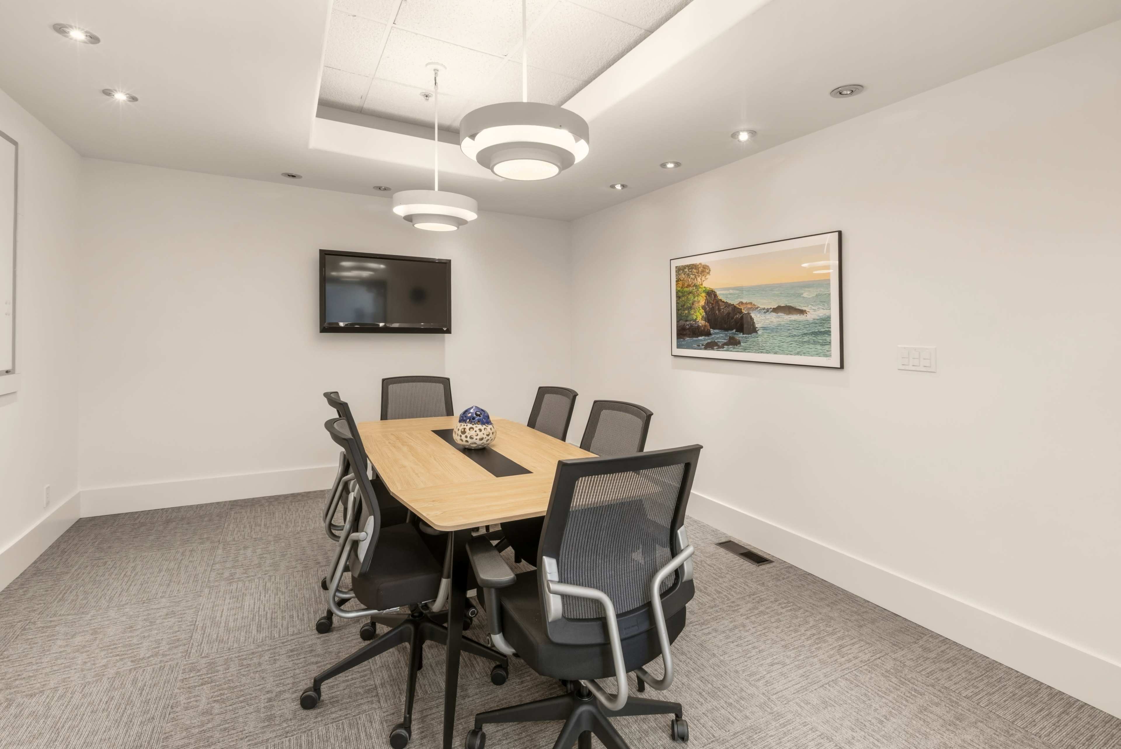 The image shows a conference room with a rectangular table surrounded by six black mesh office chairs, a television mounted on the wall, and a framed landscape picture.