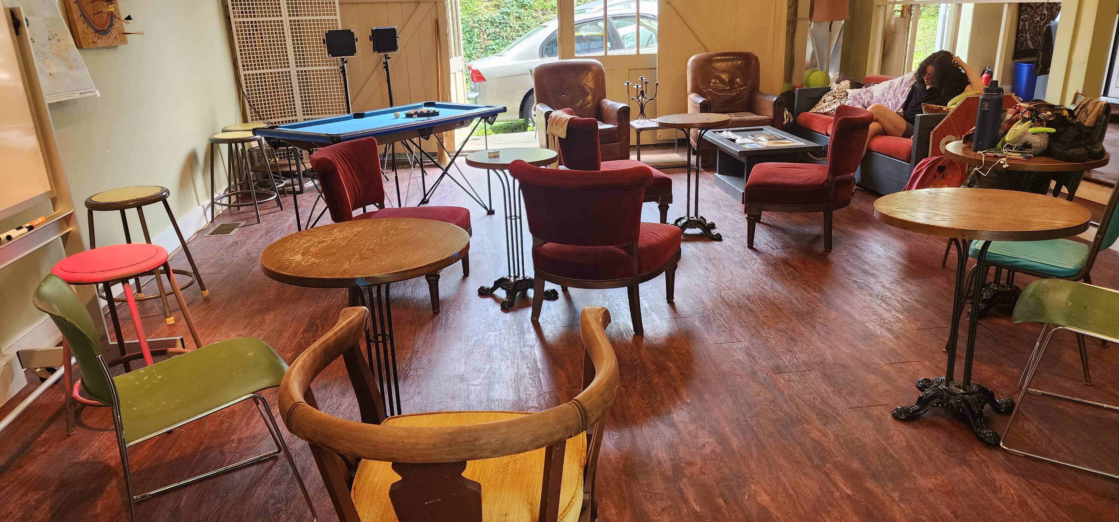 The scene shows a spacious room with several chairs and tables arranged around a blue pool table, with a couch and a car visible outside through the windows.