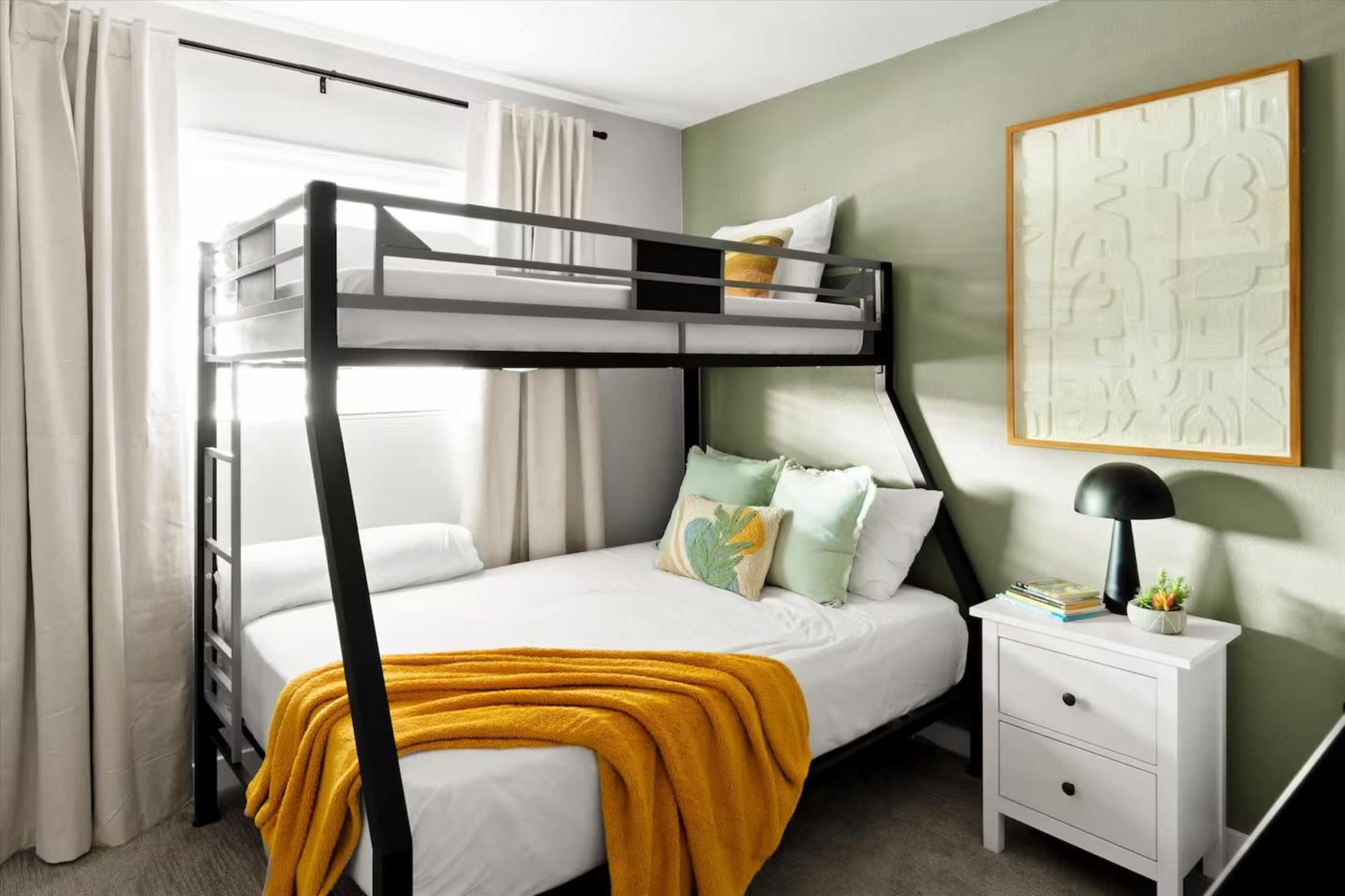 The image shows a modern bedroom featuring a black metal bunk bed, a single bed with yellow and green accents, a white nightstand, and a framed artwork on a green wall.