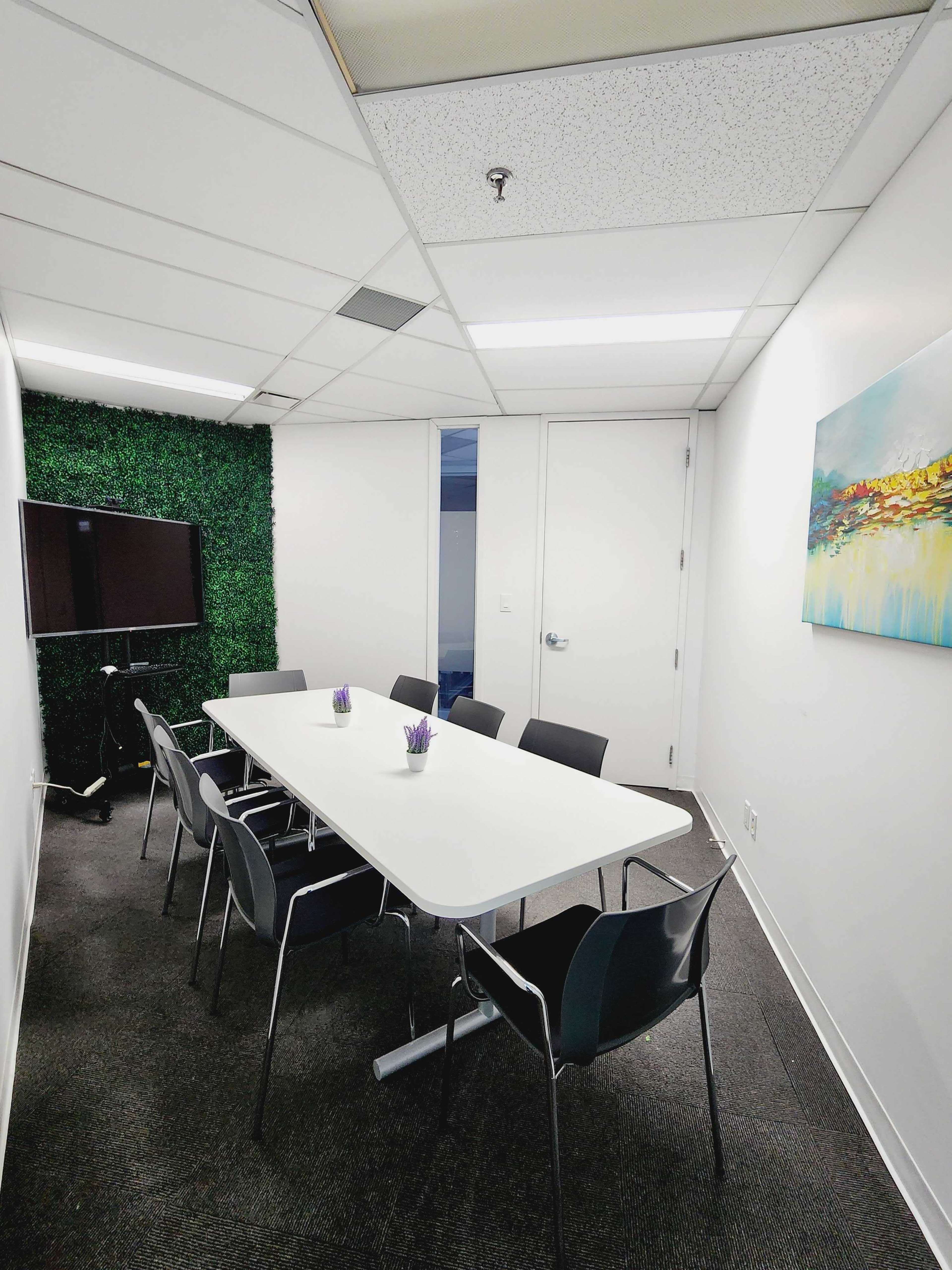 A conference room contains a white table surrounded by six chairs, a TV mounted on the wall, and decor featuring a green plant wall and a colorful painting.