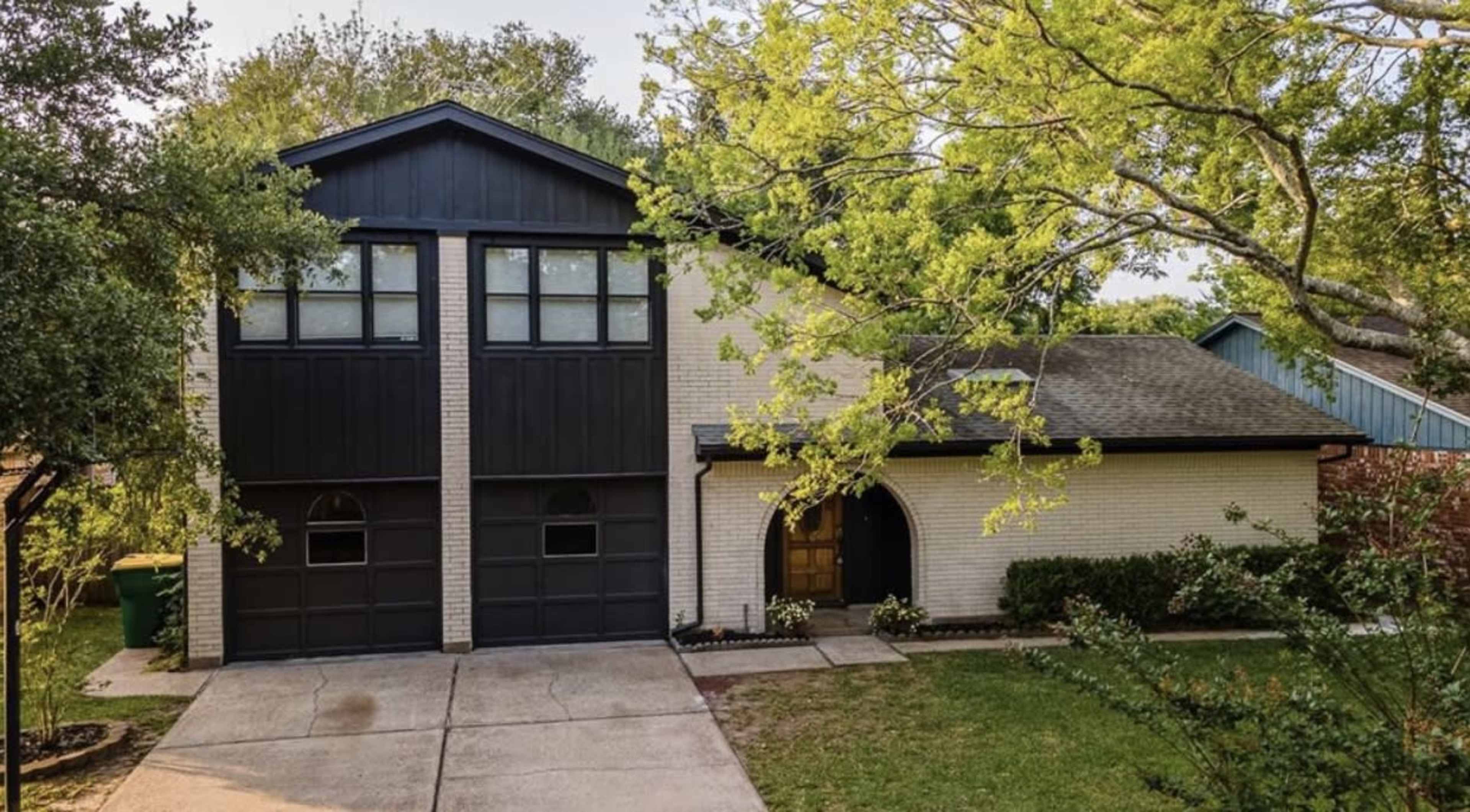 A two-story house with a black and white exterior features a double garage and a front yard with shrubs and trees.