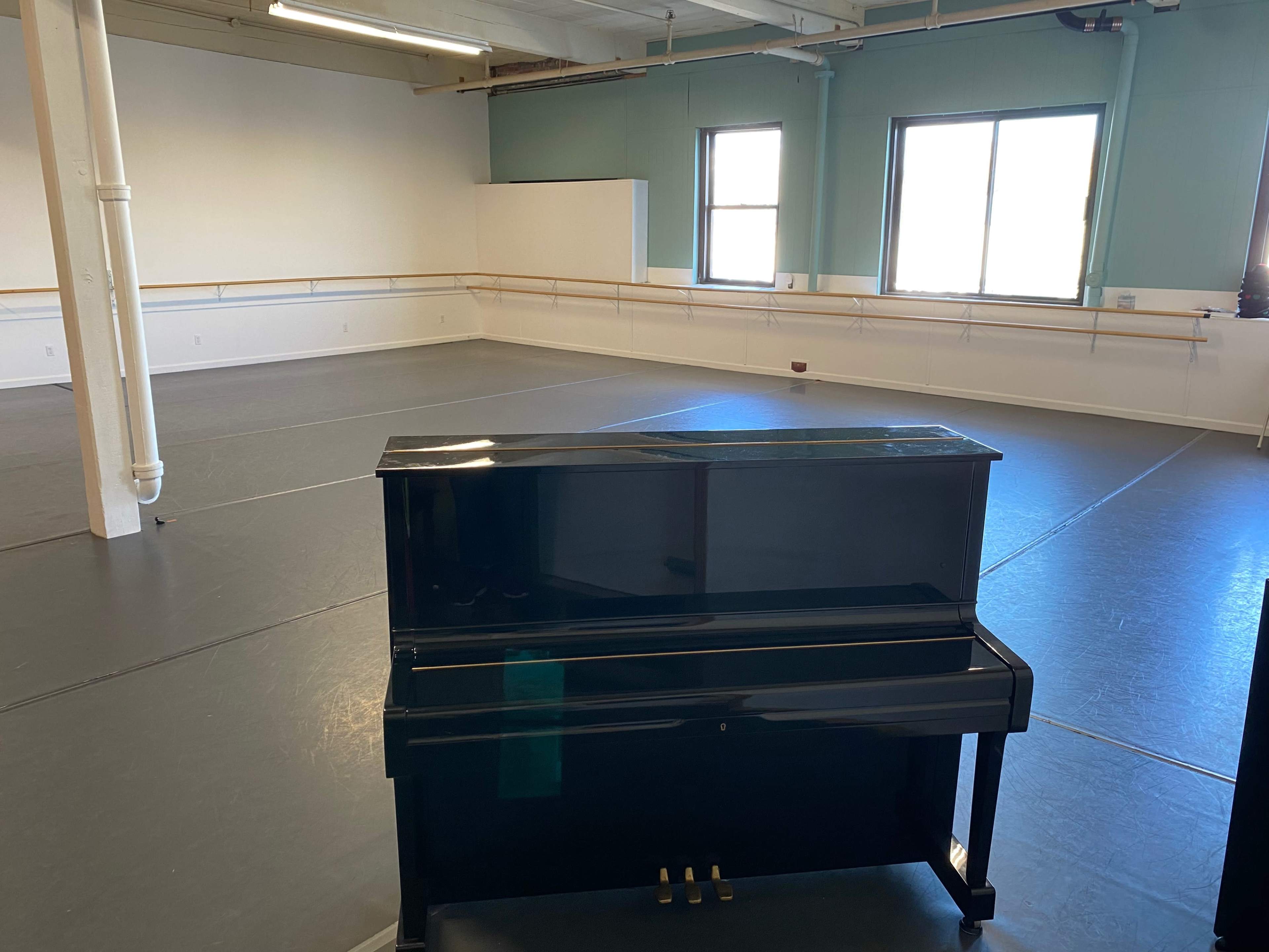 A black upright piano stands in an empty dance studio with gray flooring and large windows.