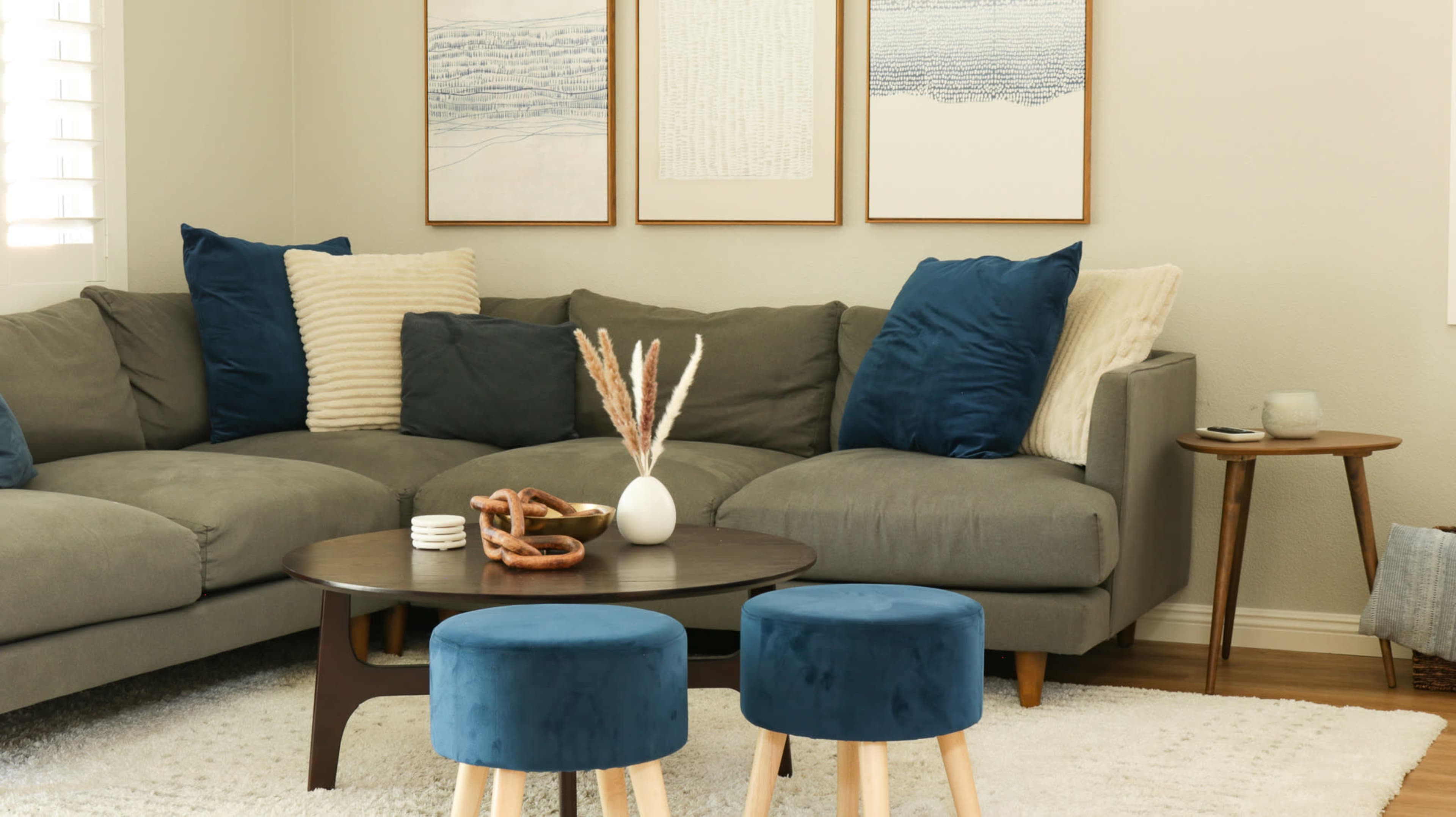 A cozy living room features a large gray sectional sofa adorned with blue and cream pillows, complemented by a round coffee table and two blue stools.