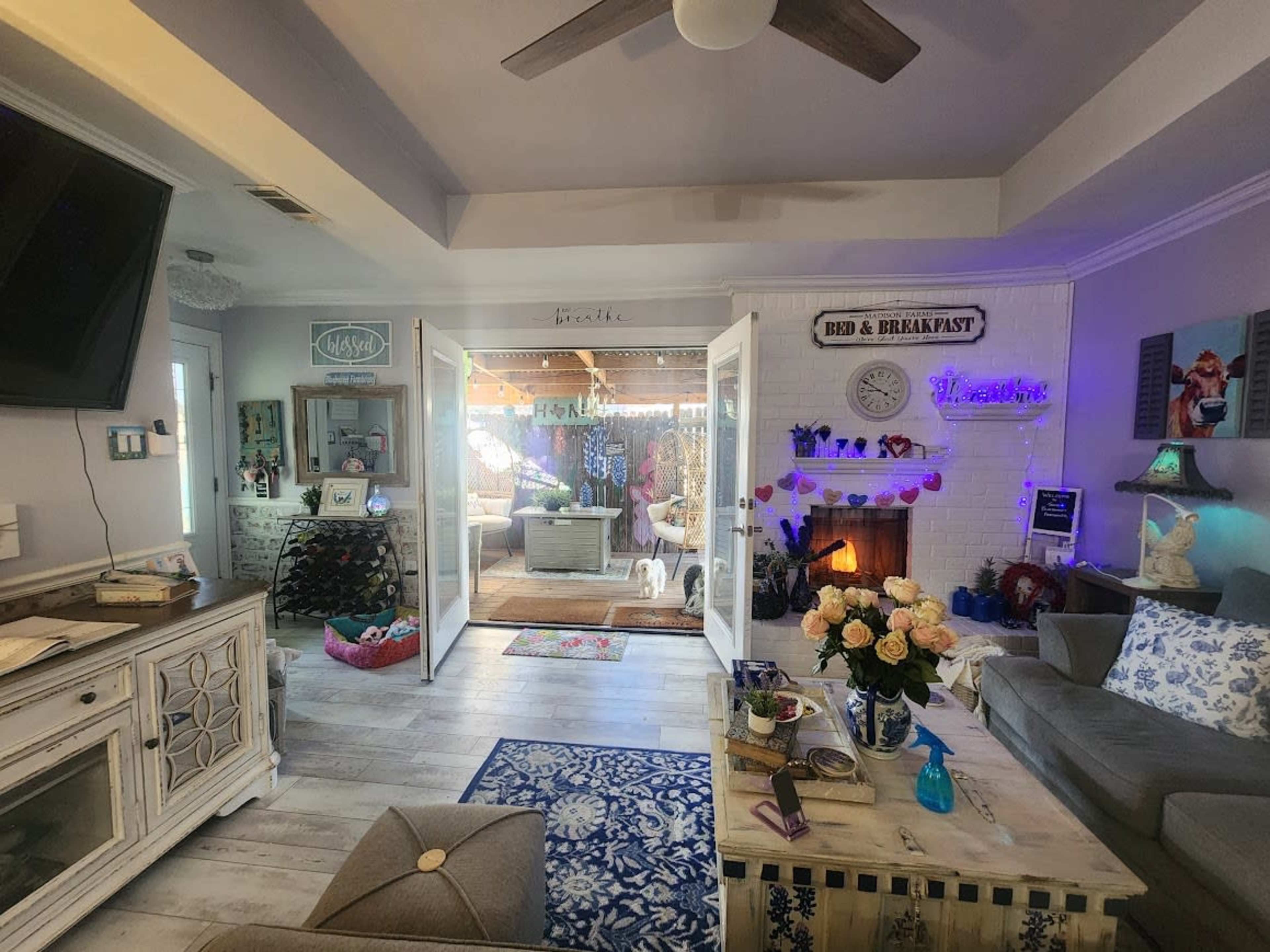 The image shows a cozy indoor space featuring a living area with a sofa, decorative plants, and an open doorway leading to a bright room with a fireplace and various decorations.
