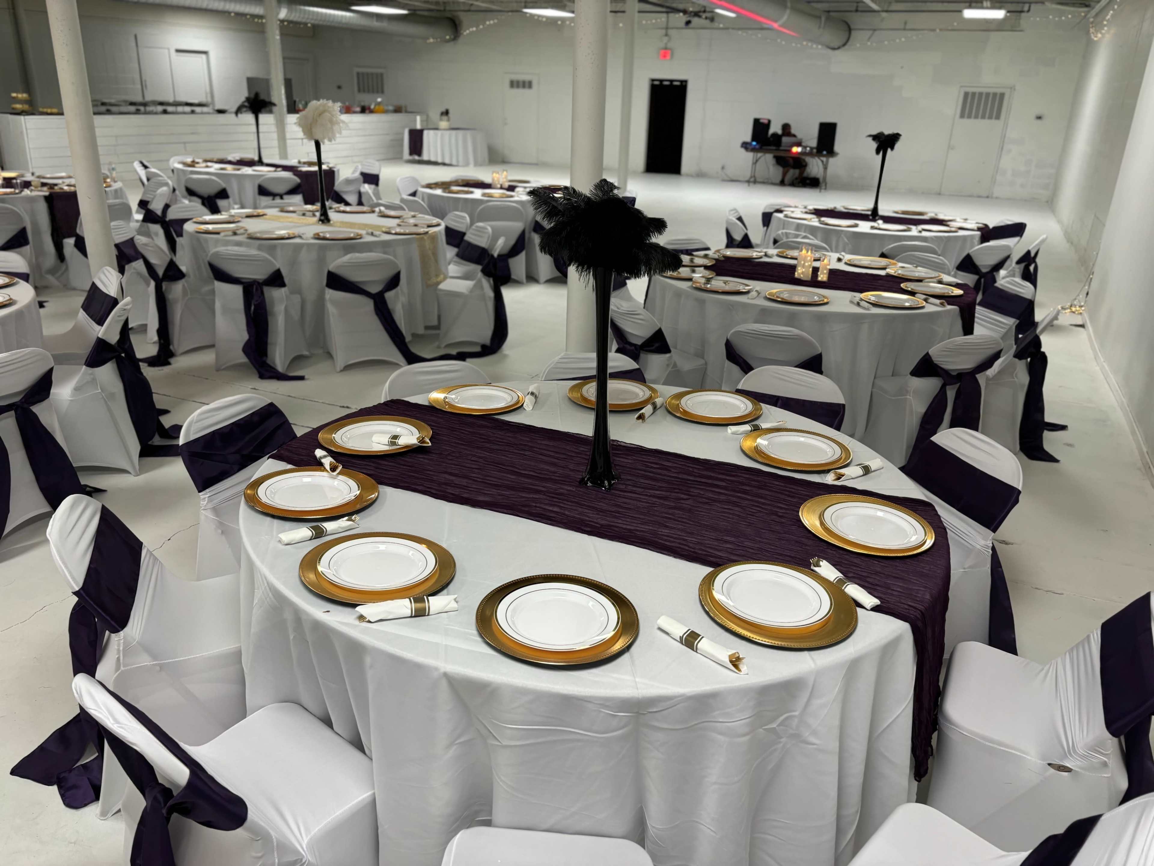 A banquet hall set with round tables covered in white tablecloths and adorned with gold-rimmed dinner plates and black feather centerpieces.