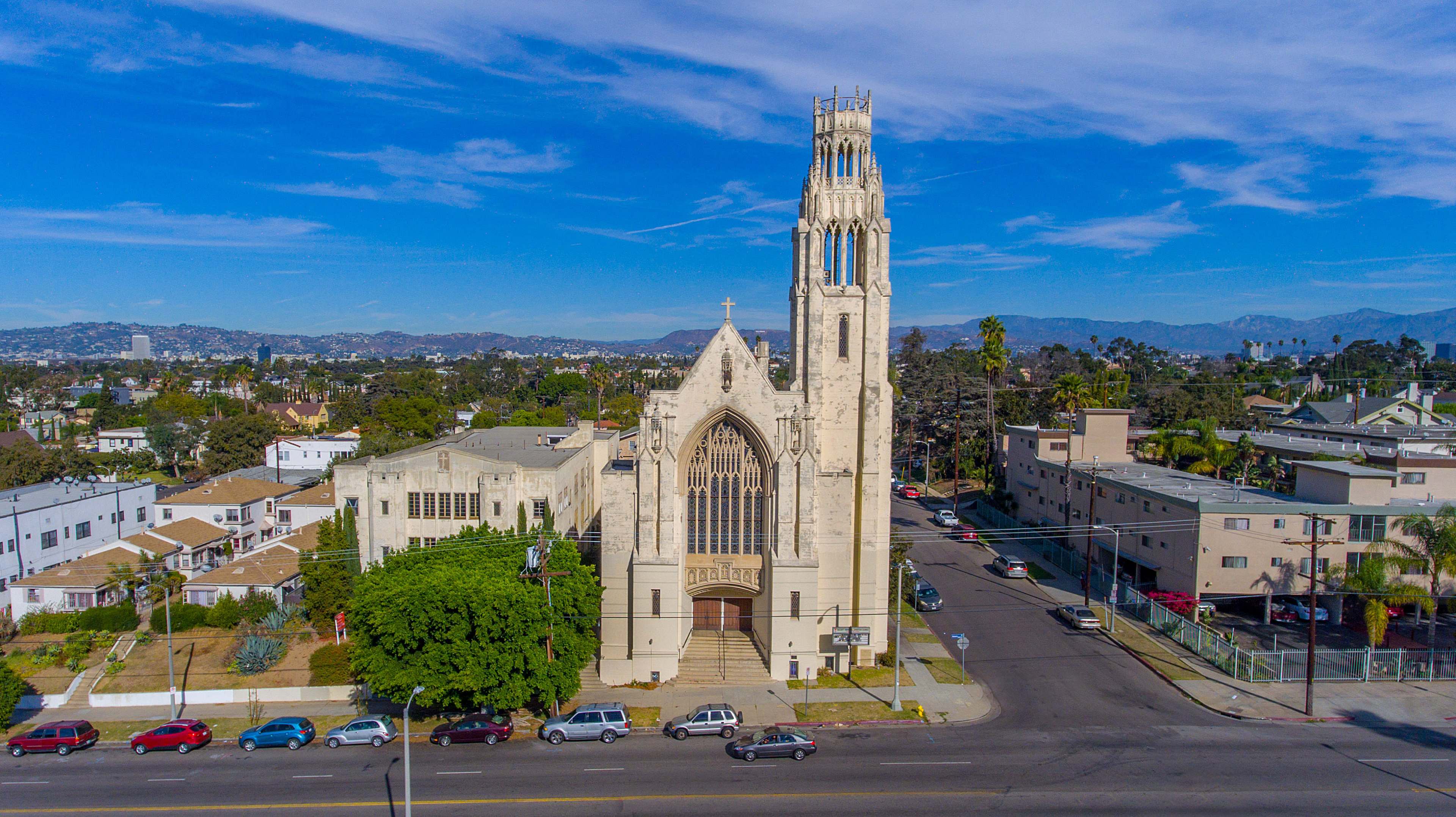 The image shows a large, ornate church building with a tall tower, located at the intersection of a street surrounded by residential and commercial buildings.