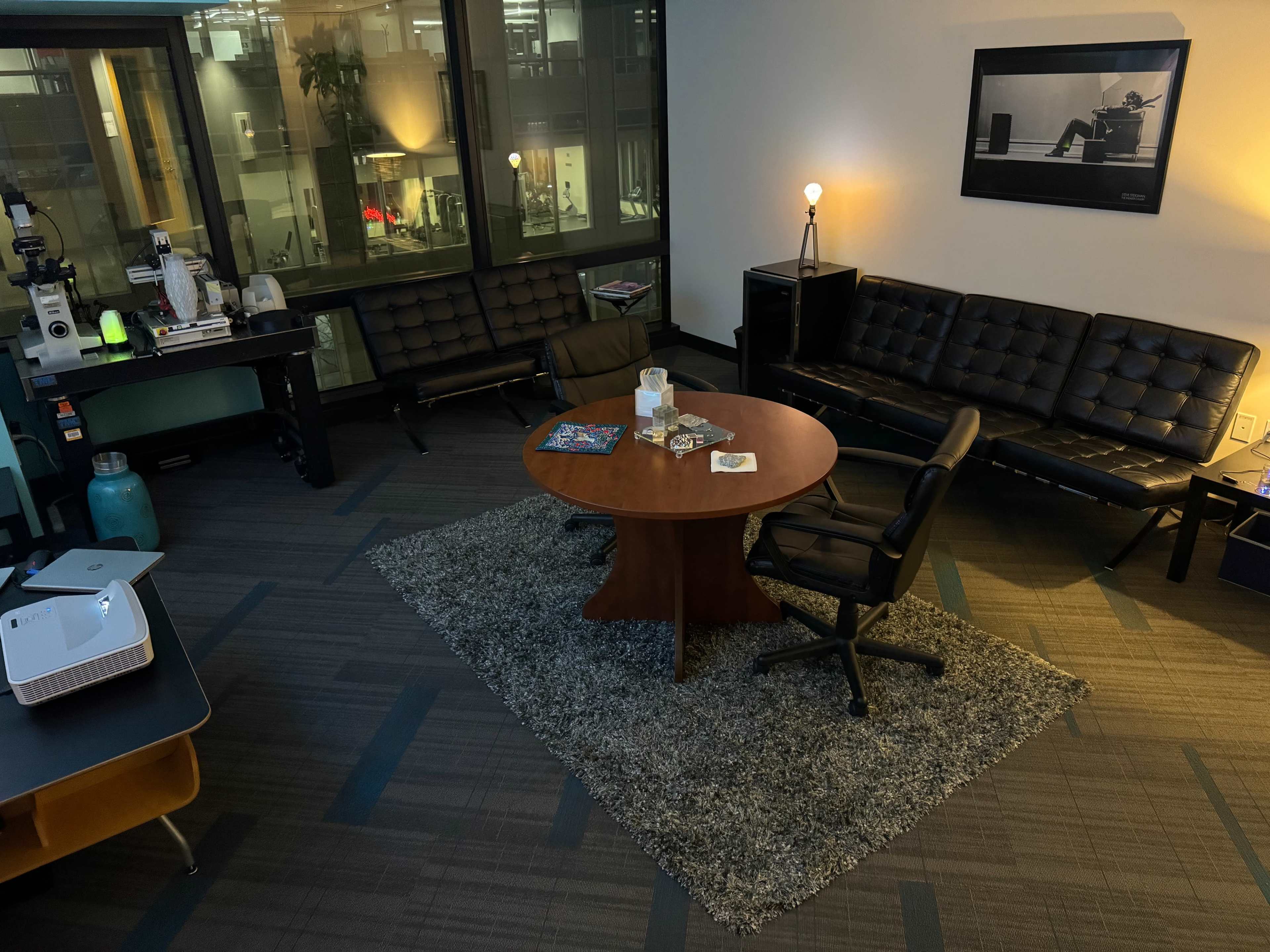 A modern office space with a round table at the center, surrounded by black seating and illuminated by a nearby lamp.