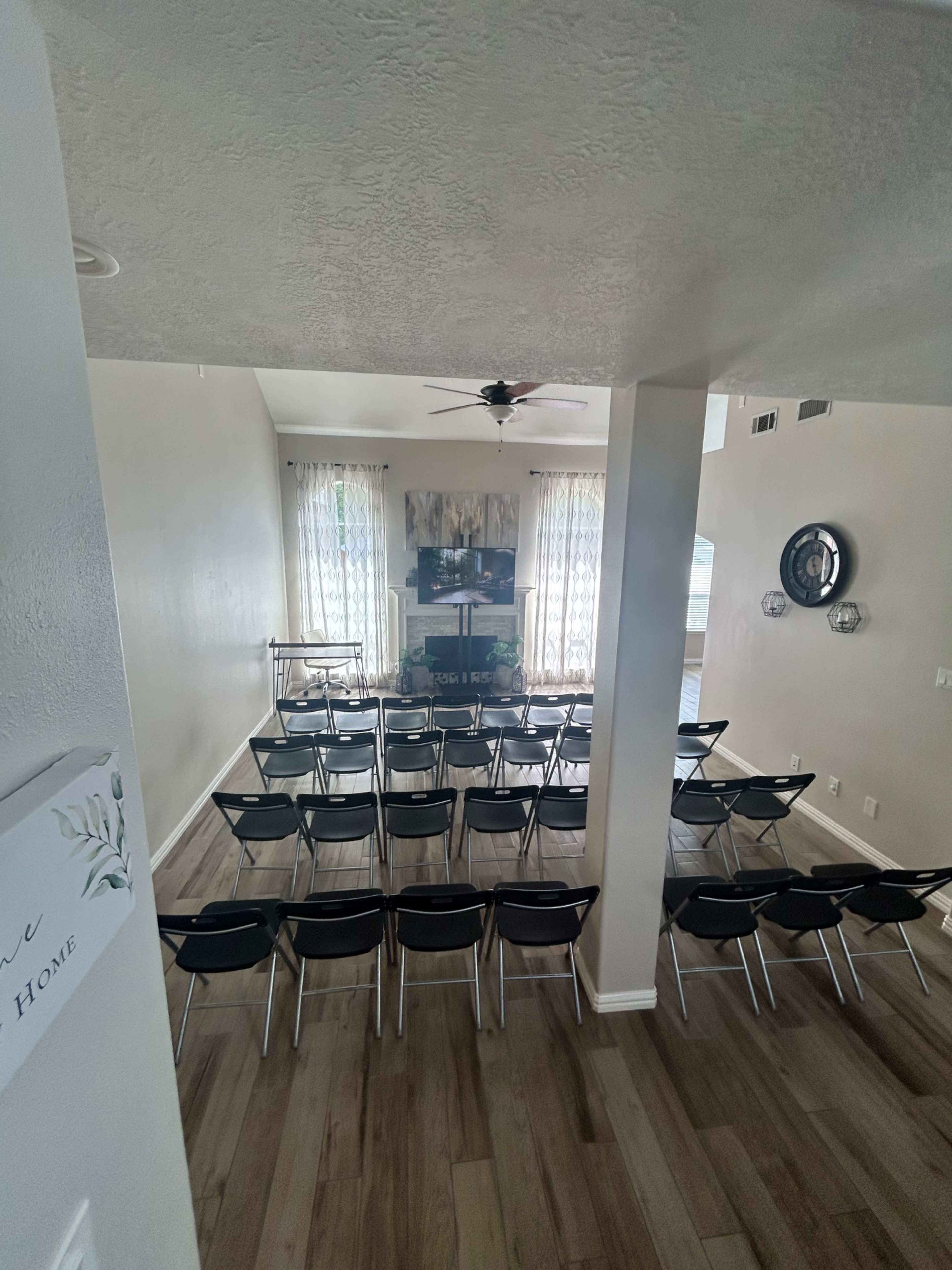 The image shows a spacious room set up with multiple black chairs arranged in rows facing a television mounted on the wall.