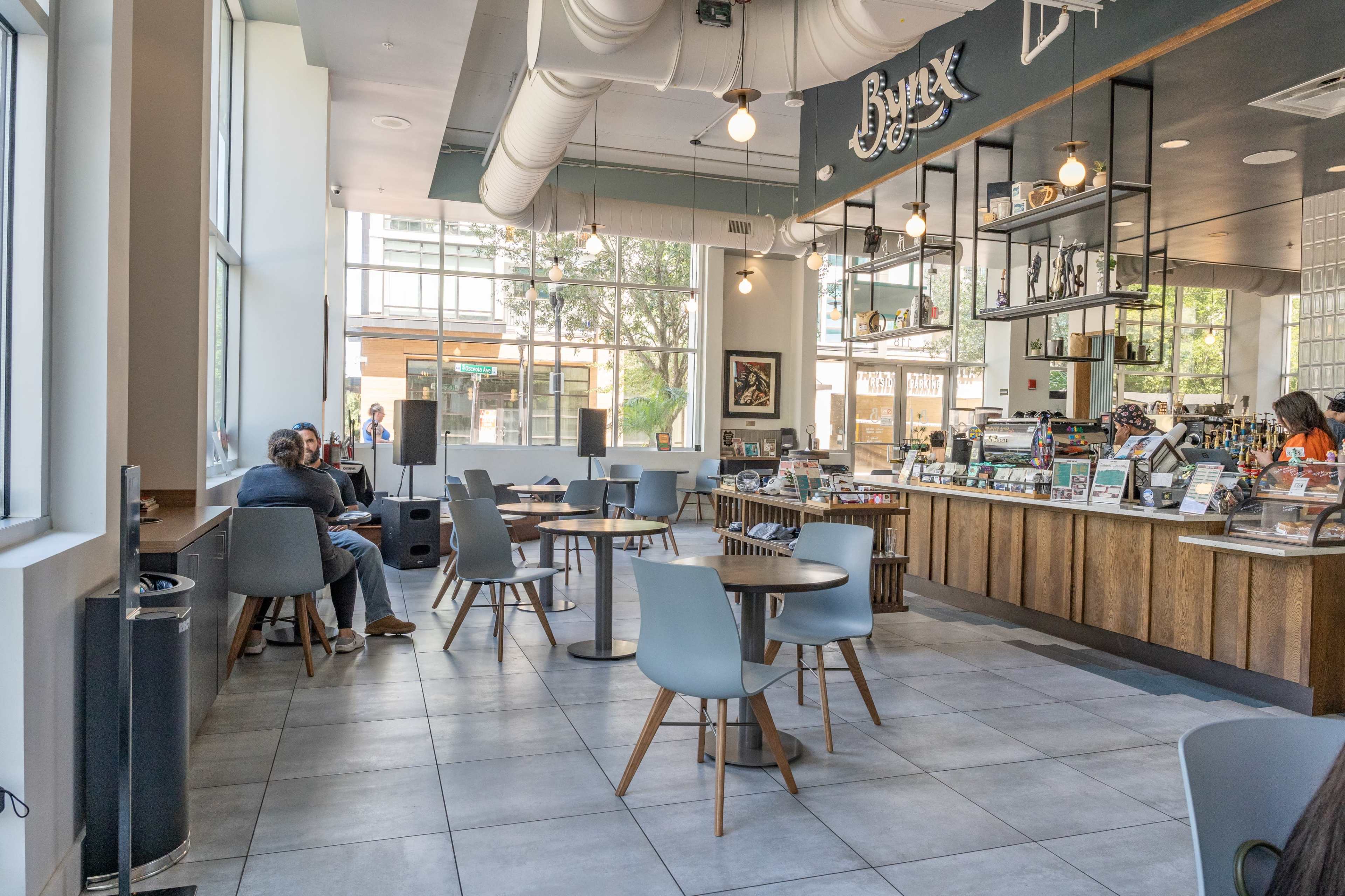 A modern café interior with wooden counters, gray tables and chairs, and large windows allowing natural light to enter.
