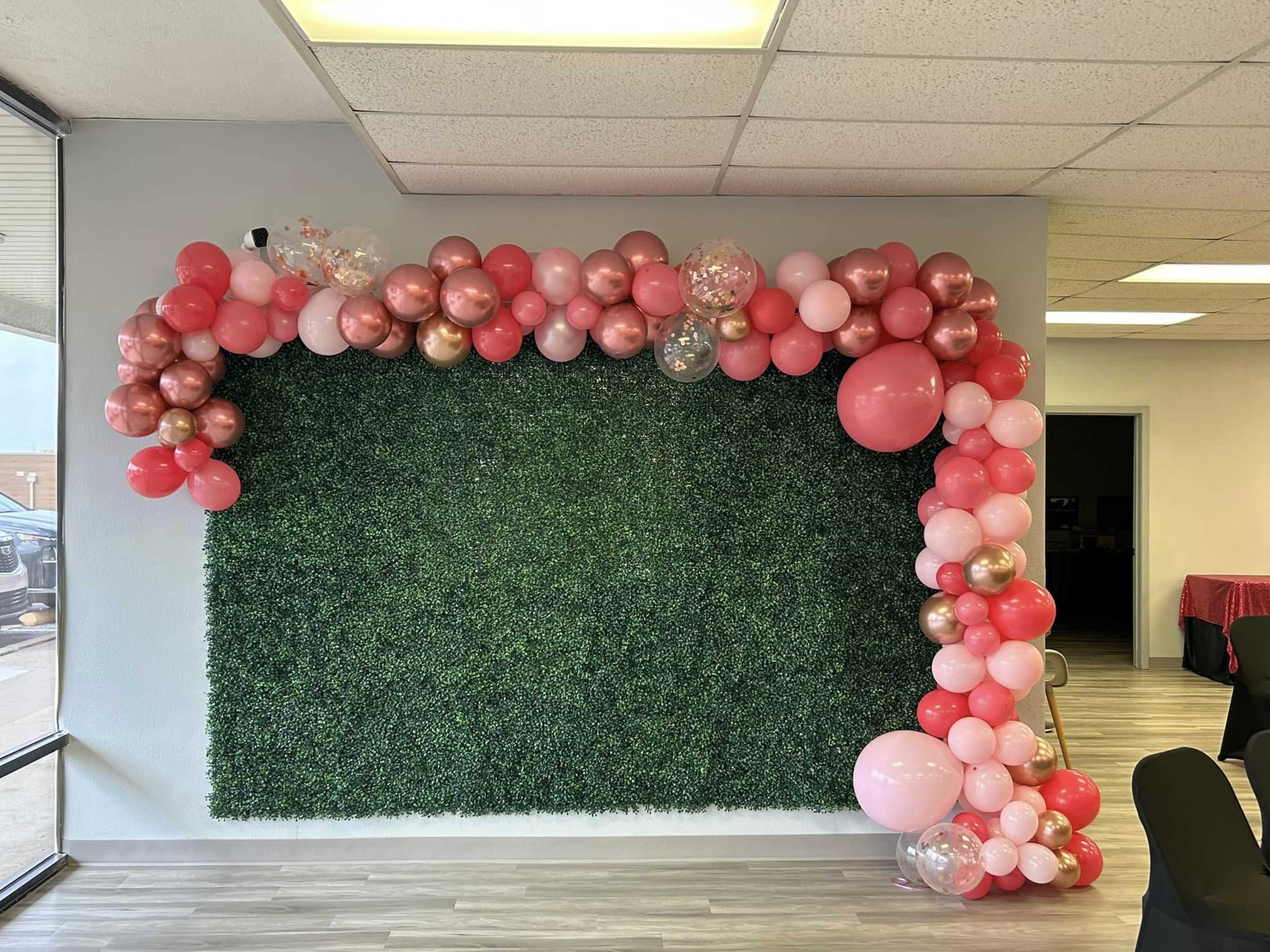 A wall adorned with a floral arrangement of pink, red, and gold balloons is set against a green artificial shrub backdrop.