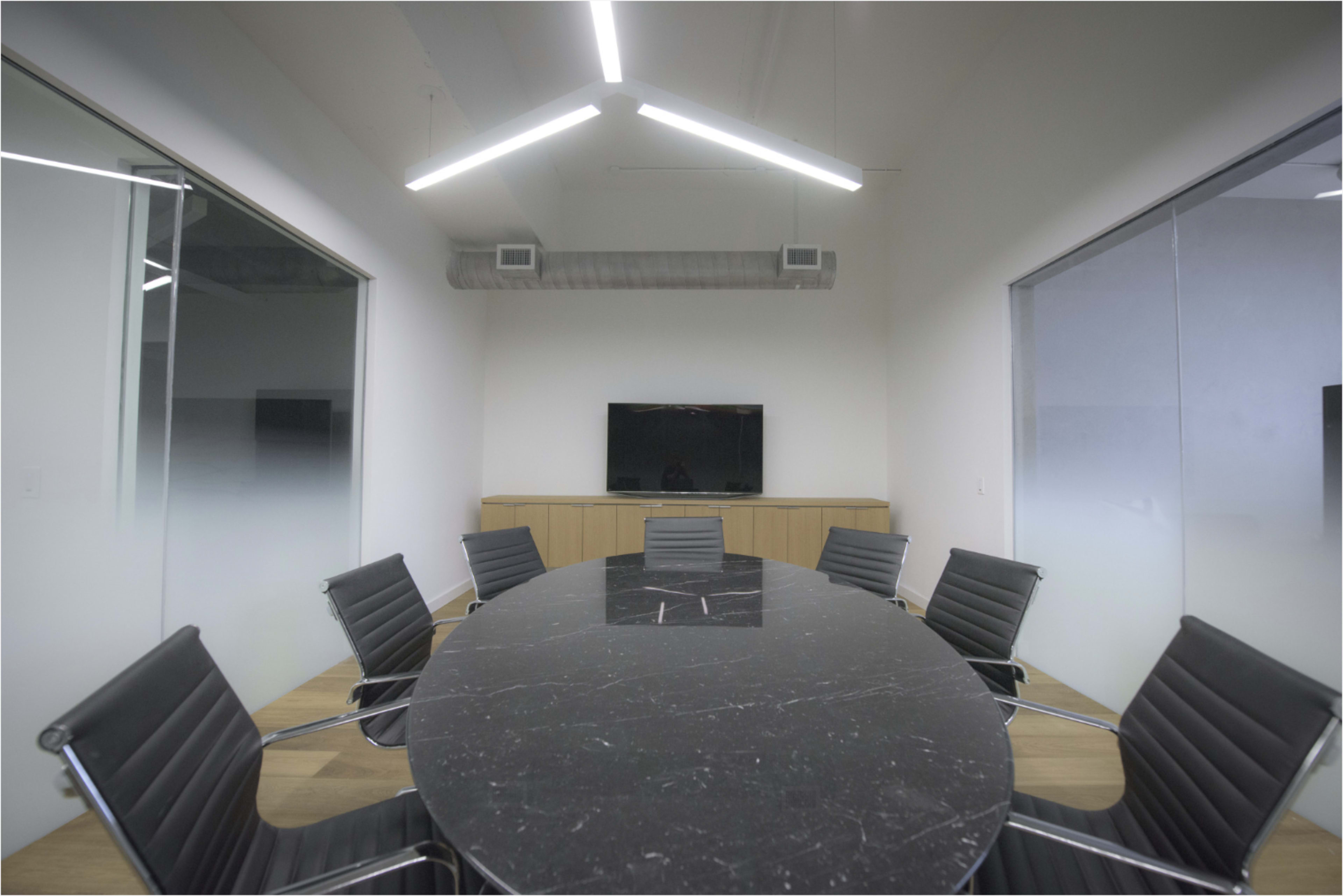 The image shows a modern conference room with a large black oval table, eight black chairs, and a wall-mounted television on a wooden cabinet.