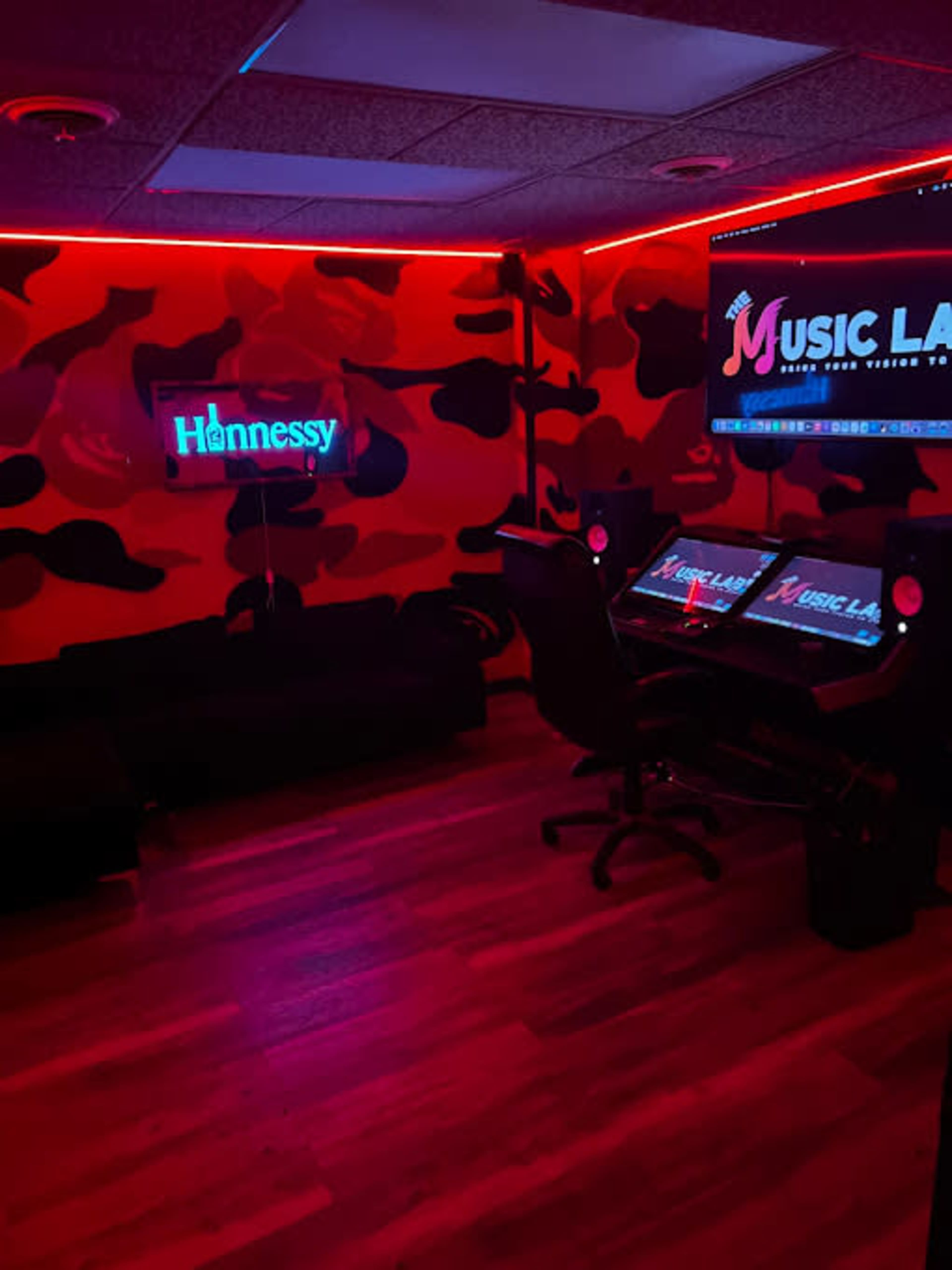 A room with red lighting, a camouflage-patterned wall, a sofa, and a desk with computers displaying music-related content.