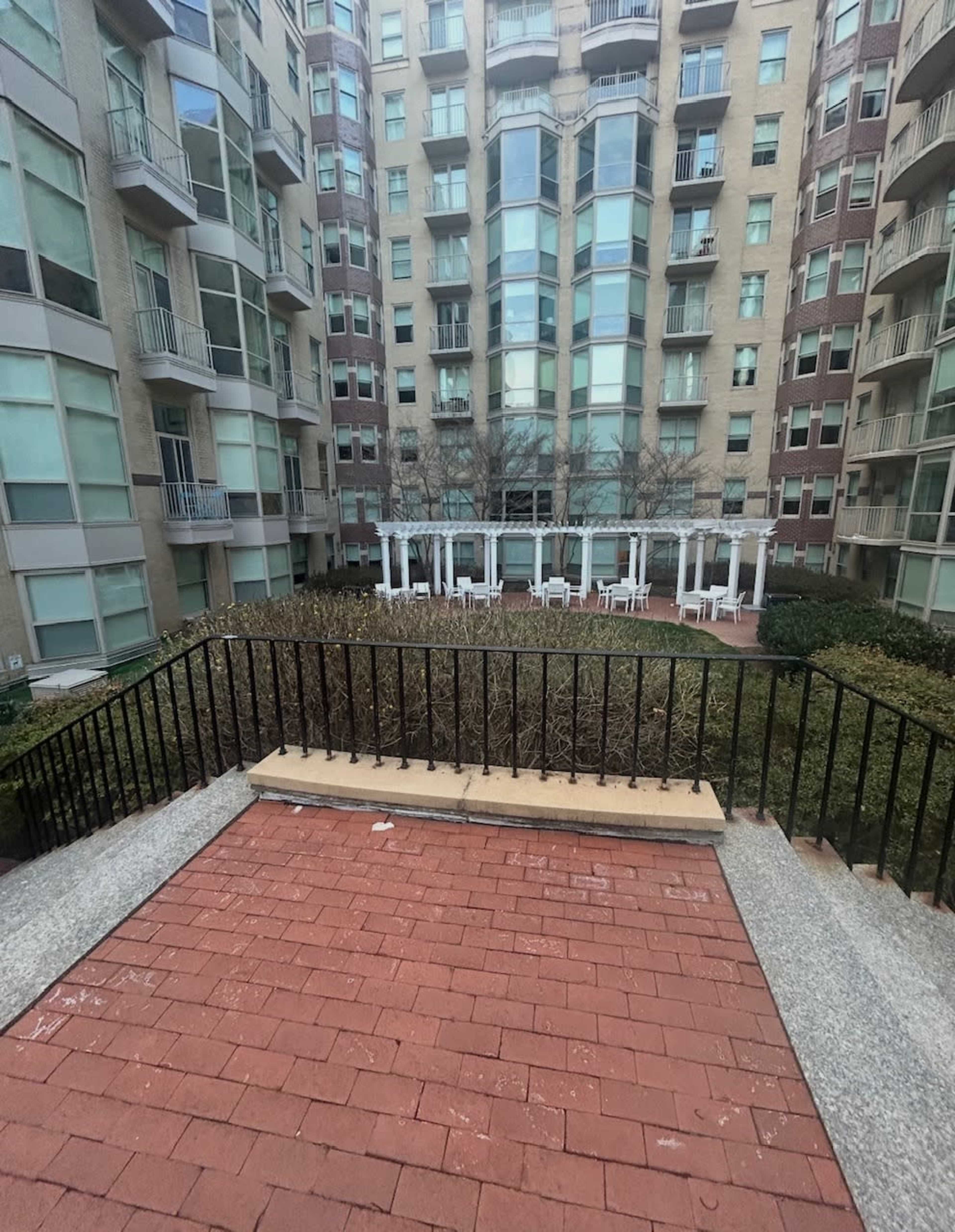 The image shows a brick walkway leading to a landscaped courtyard surrounded by several multi-story apartment buildings.