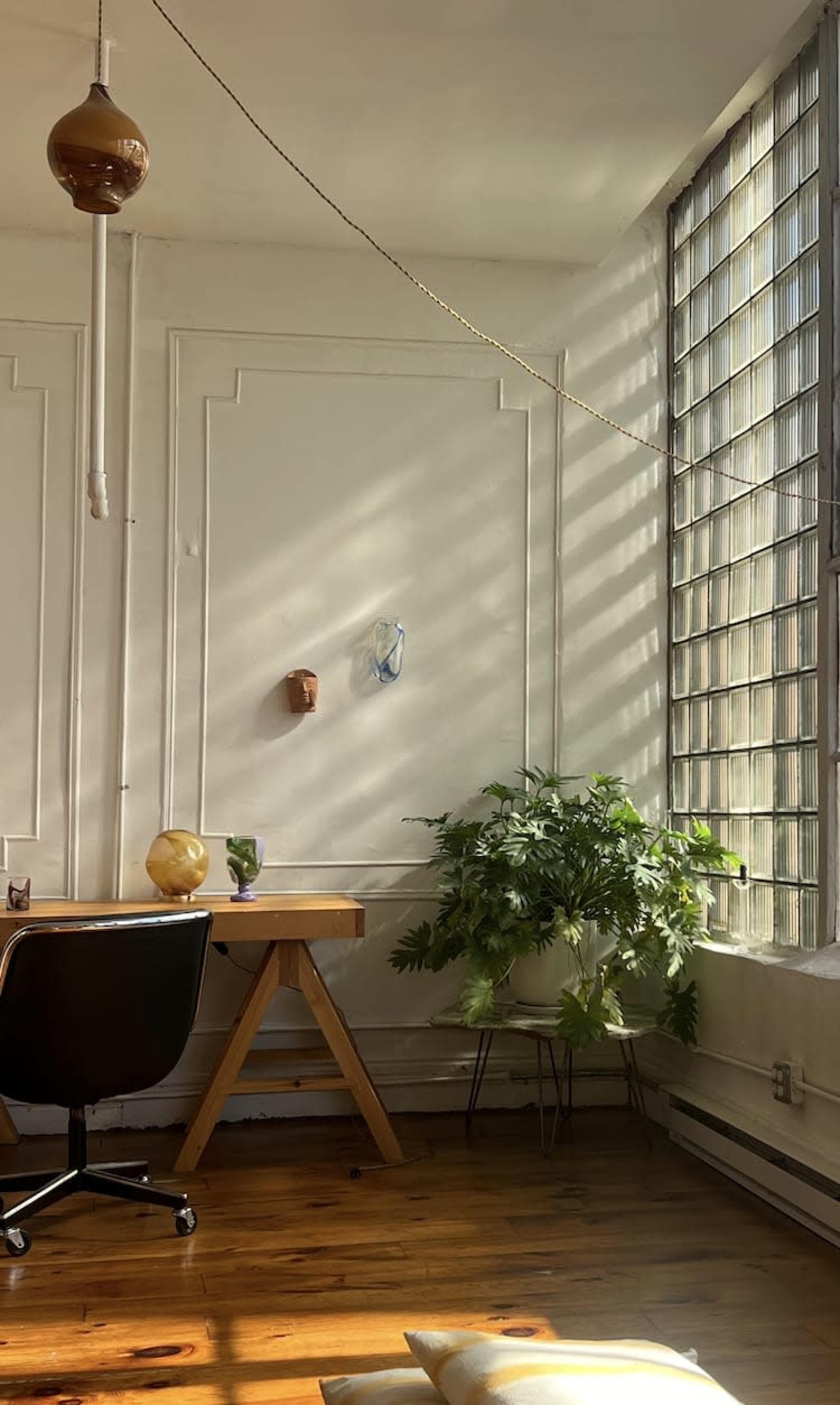 The image shows a well-lit corner of a room featuring a wooden desk with a black chair, a plant beside a large window, and decorative wall elements.
