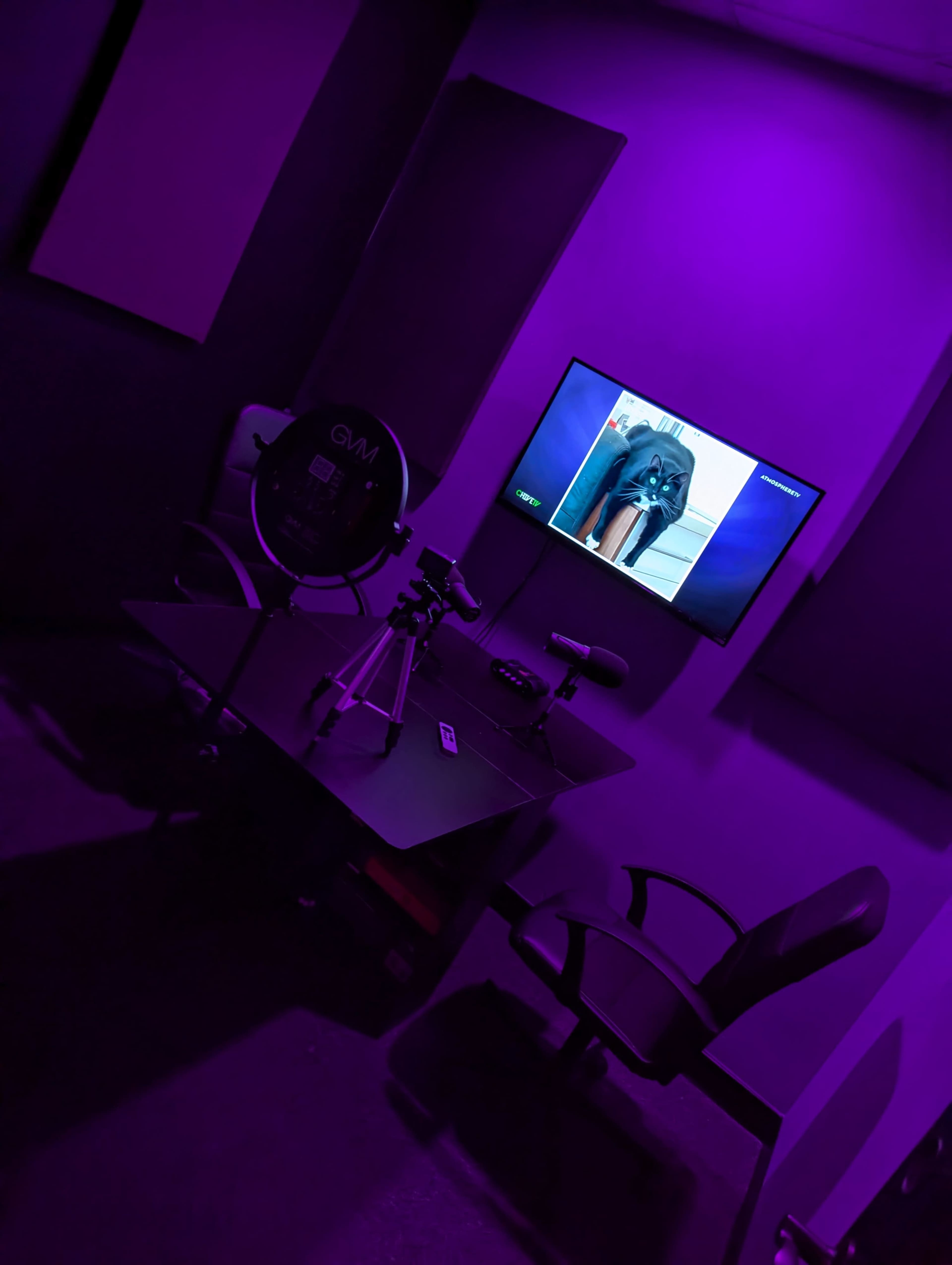 A dimly lit room features a black table with a camera on a tripod, a microphone, and a wall-mounted television displaying an image.