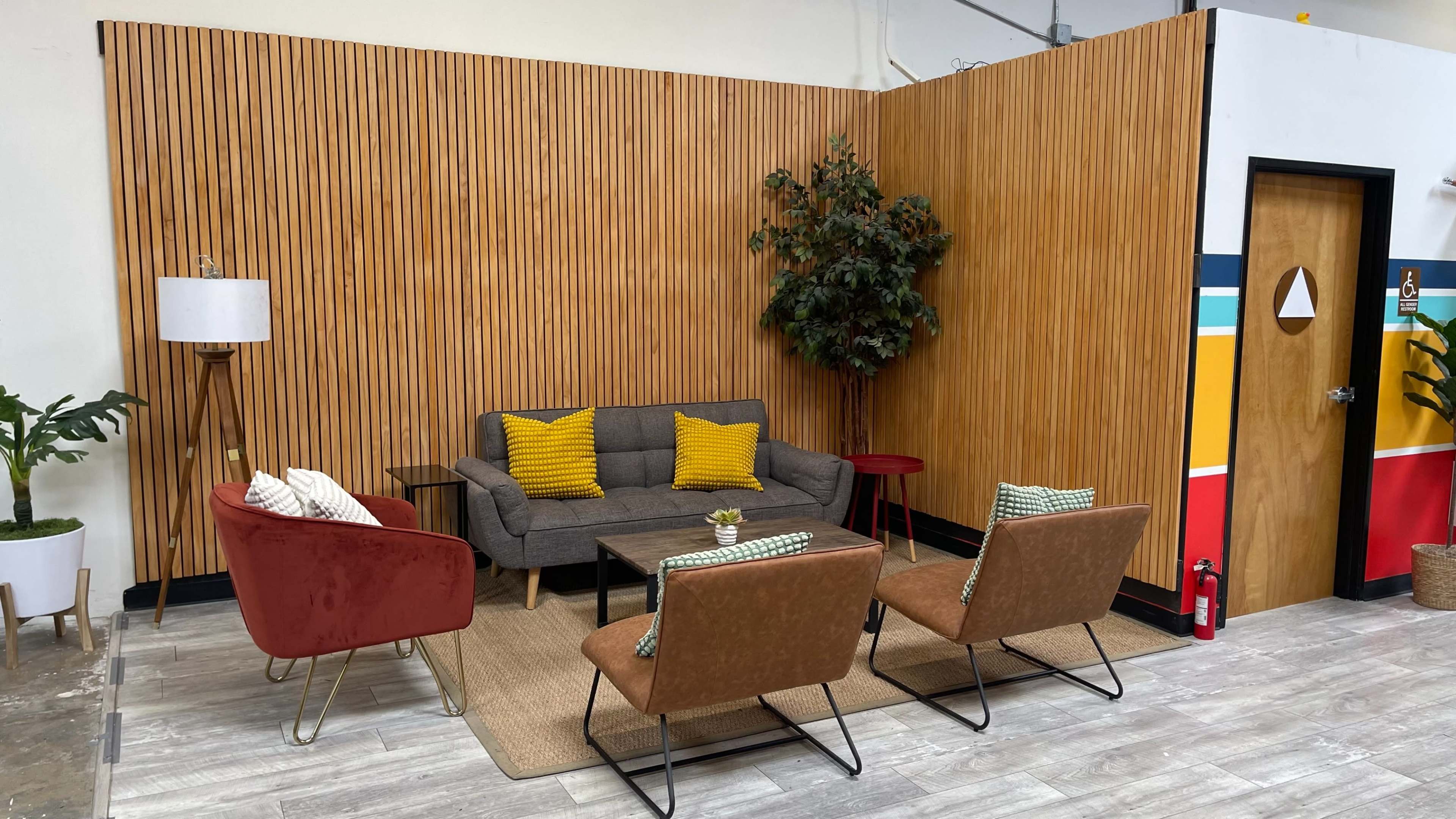 A modern seating area with a gray sofa, two brown chairs, a small table, and potted plants against a wooden wall.