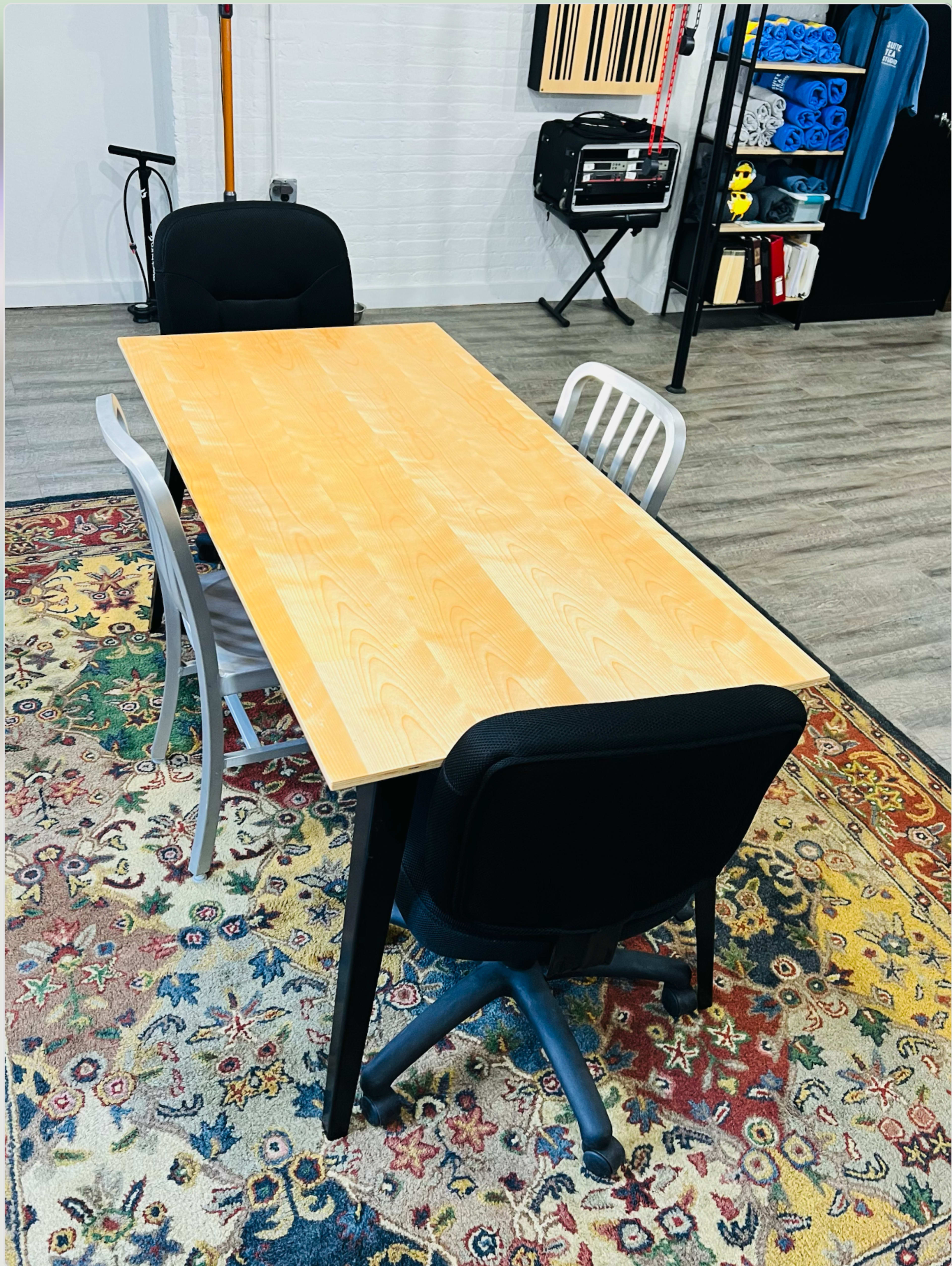 A rectangular wooden table is surrounded by three chairs on a patterned area rug in a room with various equipment in the background.