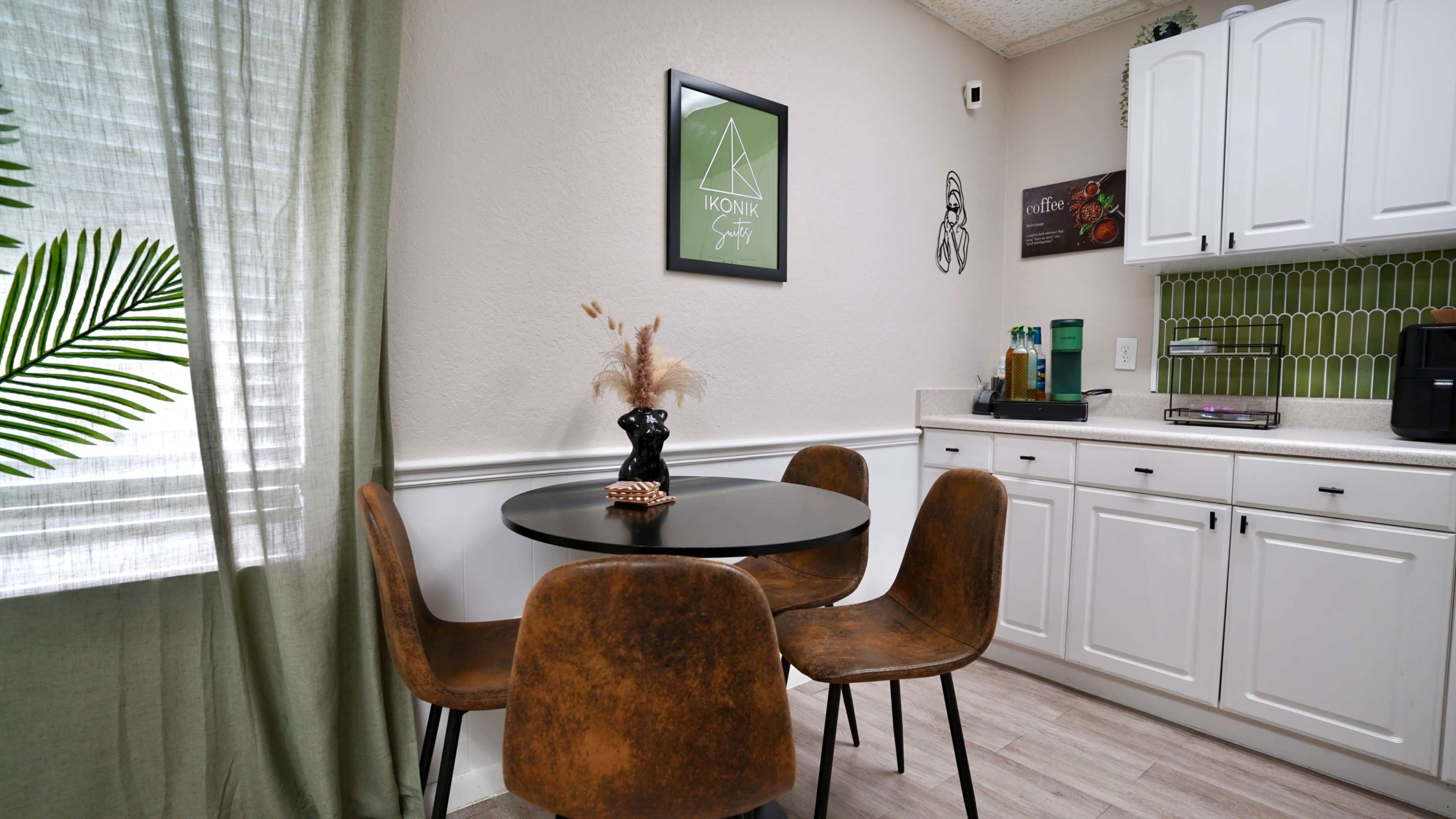 A small dining area features a round black table surrounded by four brown chairs, with a potted plant and decorative elements on the table against a light-colored wall.