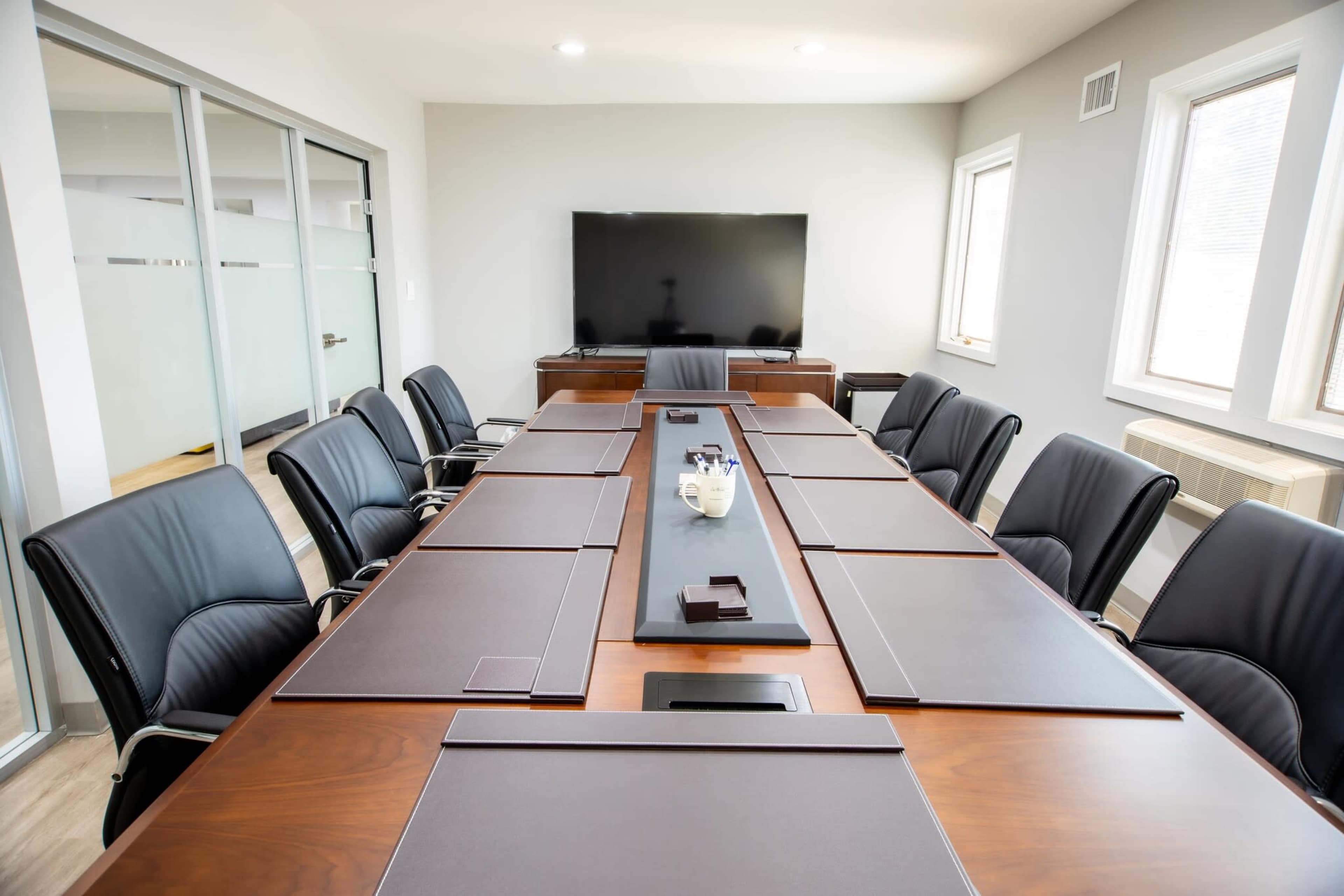 A modern conference room features a long wooden table surrounded by black leather chairs, with a large screen on the wall and natural light coming through windows.