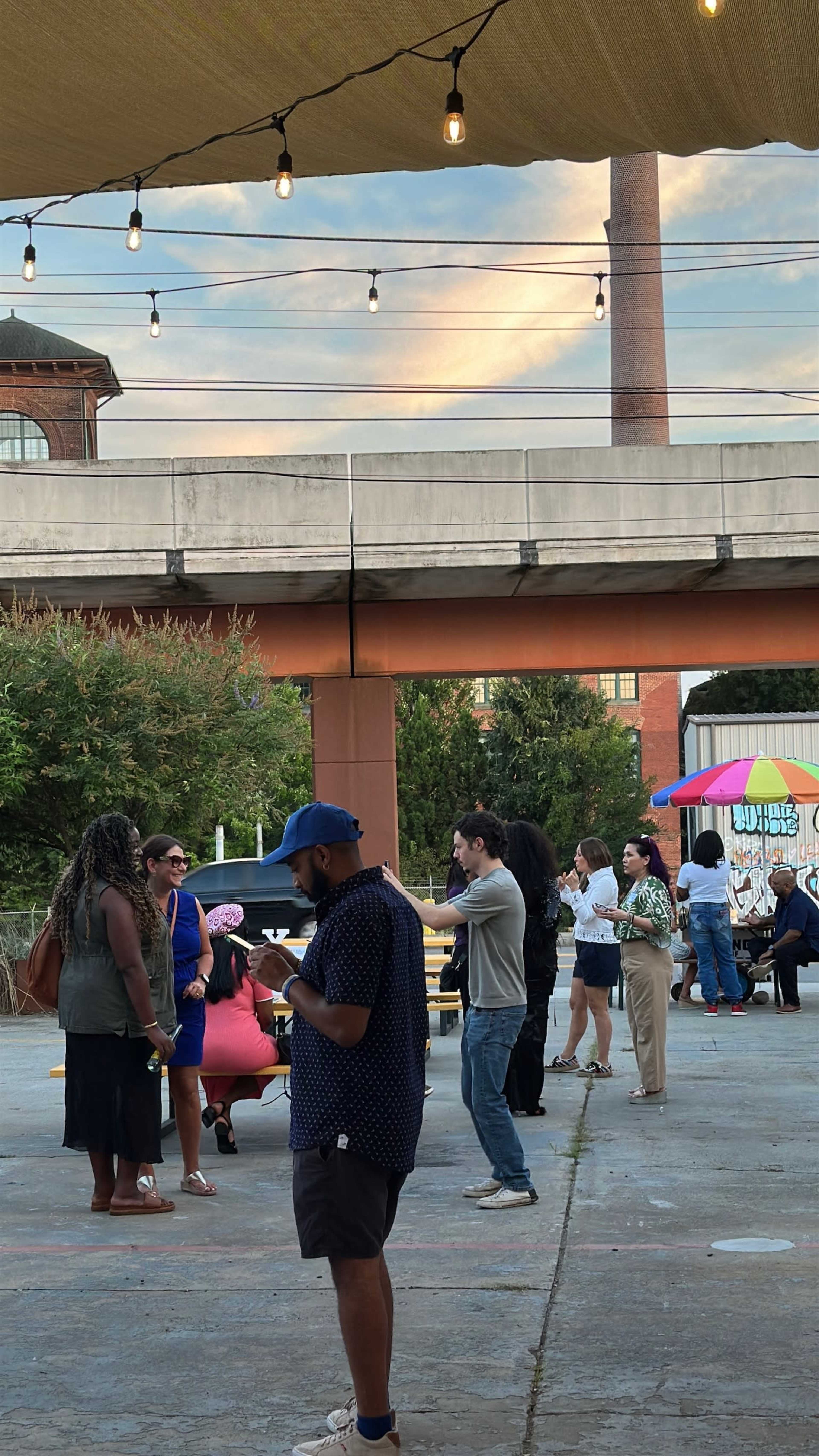 The image shows a gathering of people socializing in an outdoor space with string lights overhead and a building in the background.