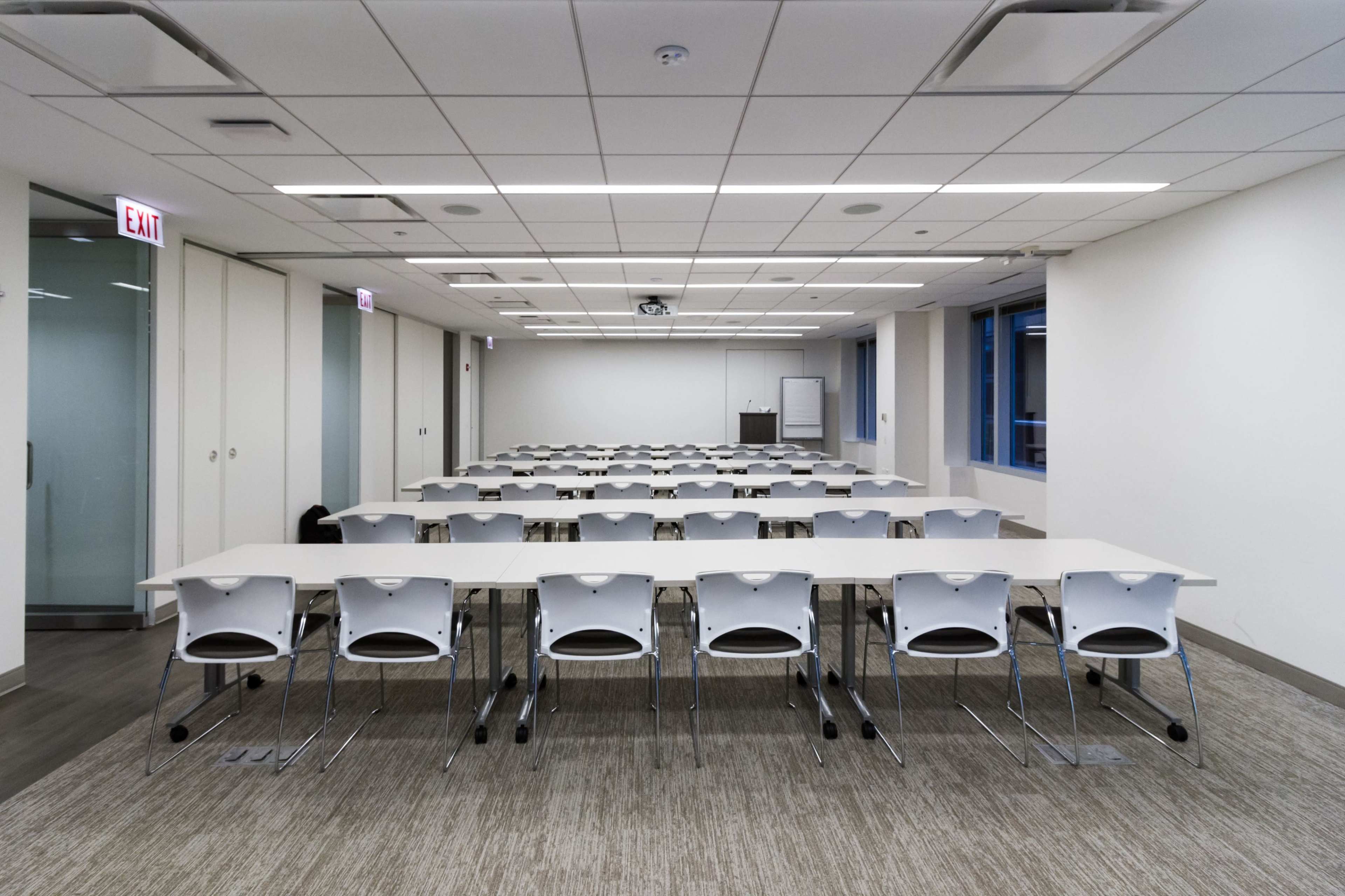 A modern meeting room features rows of tables and chairs arranged for a workshop or presentation, with large windows and a projector screen at one end.