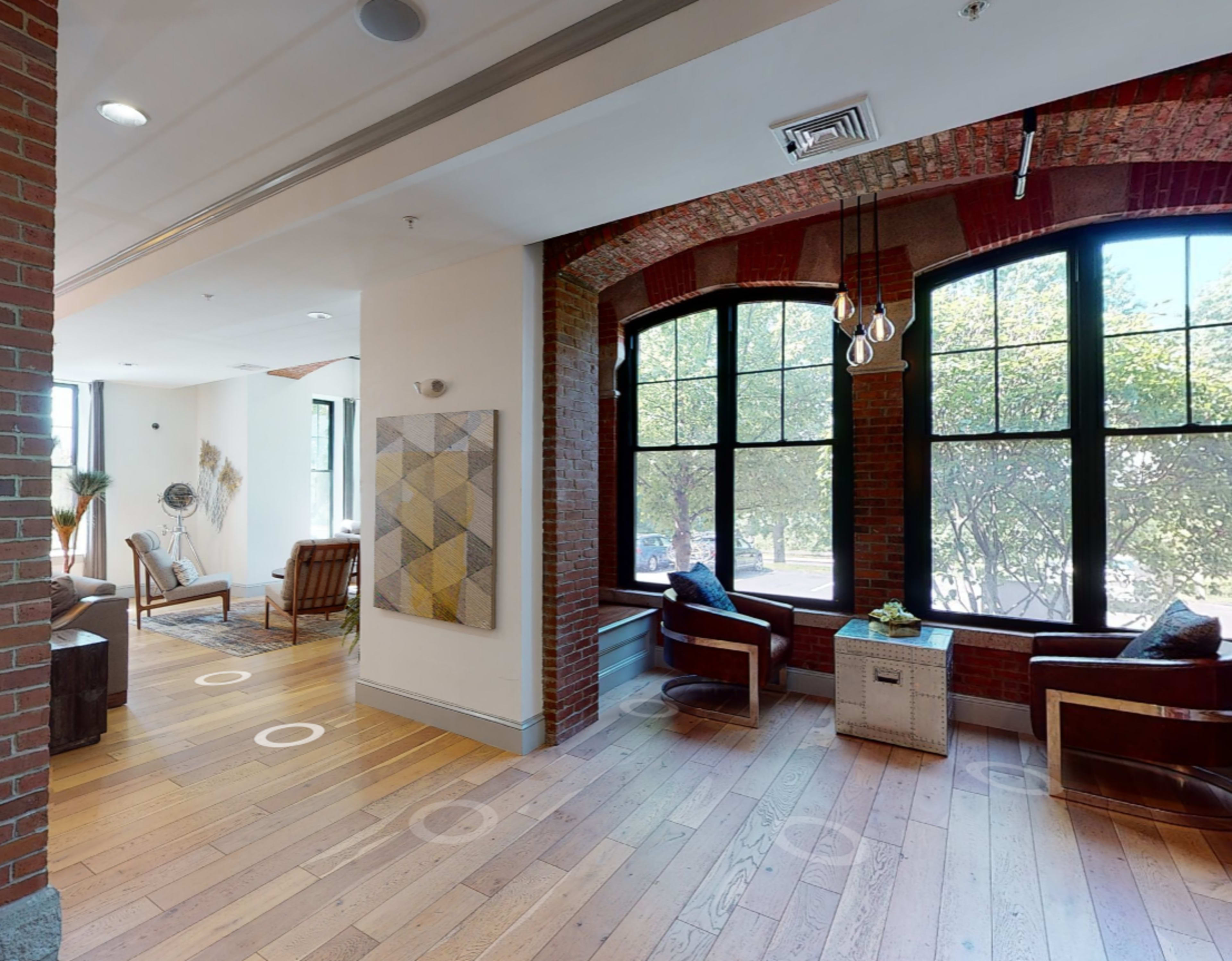 A brightly lit room features large windows, hardwood floors, and various seating areas, including two chairs and artwork on the walls.