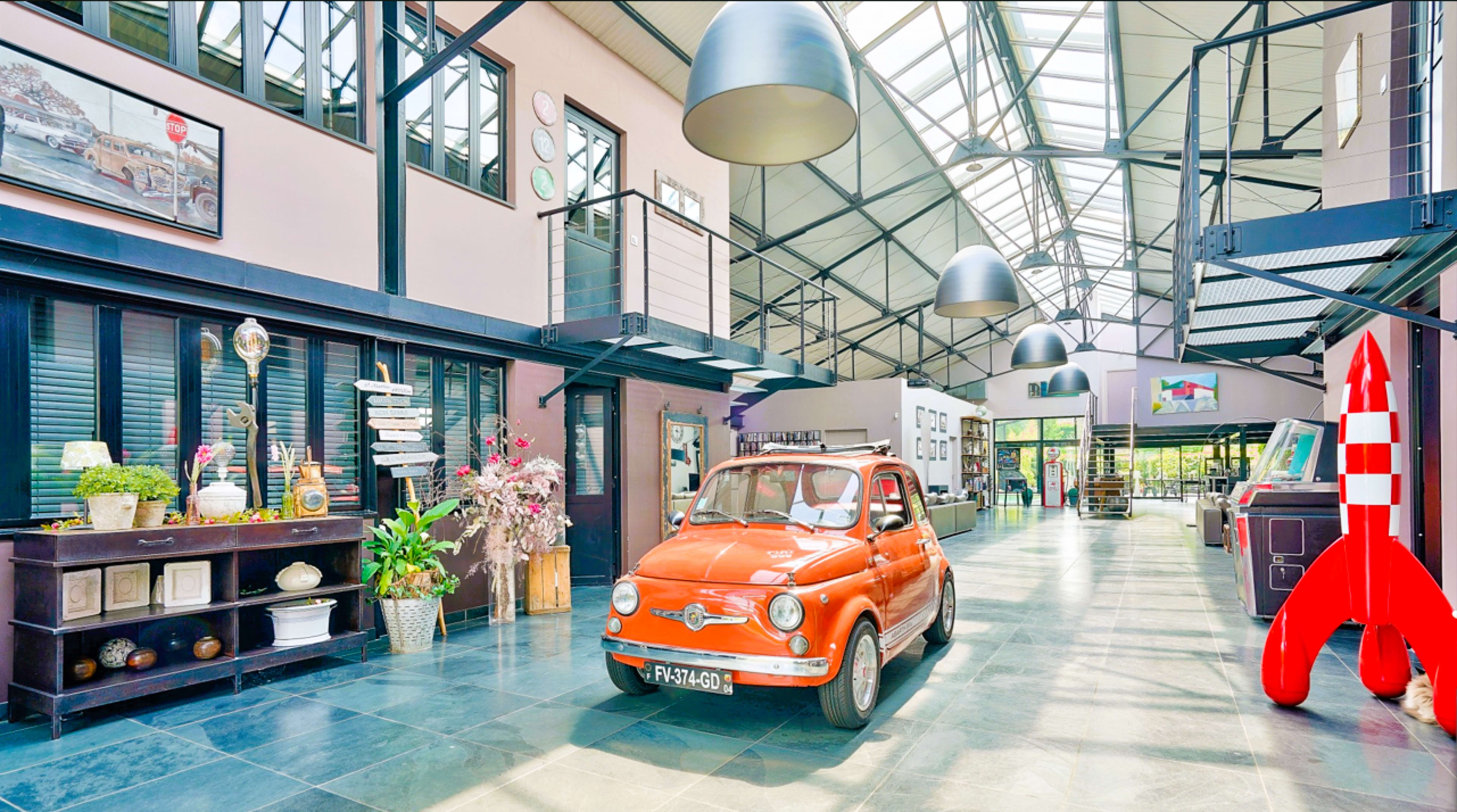 An orange vintage car is positioned in a spacious, modern industrial-style interior with high ceilings and large windows, featuring decorative artwork and plants.