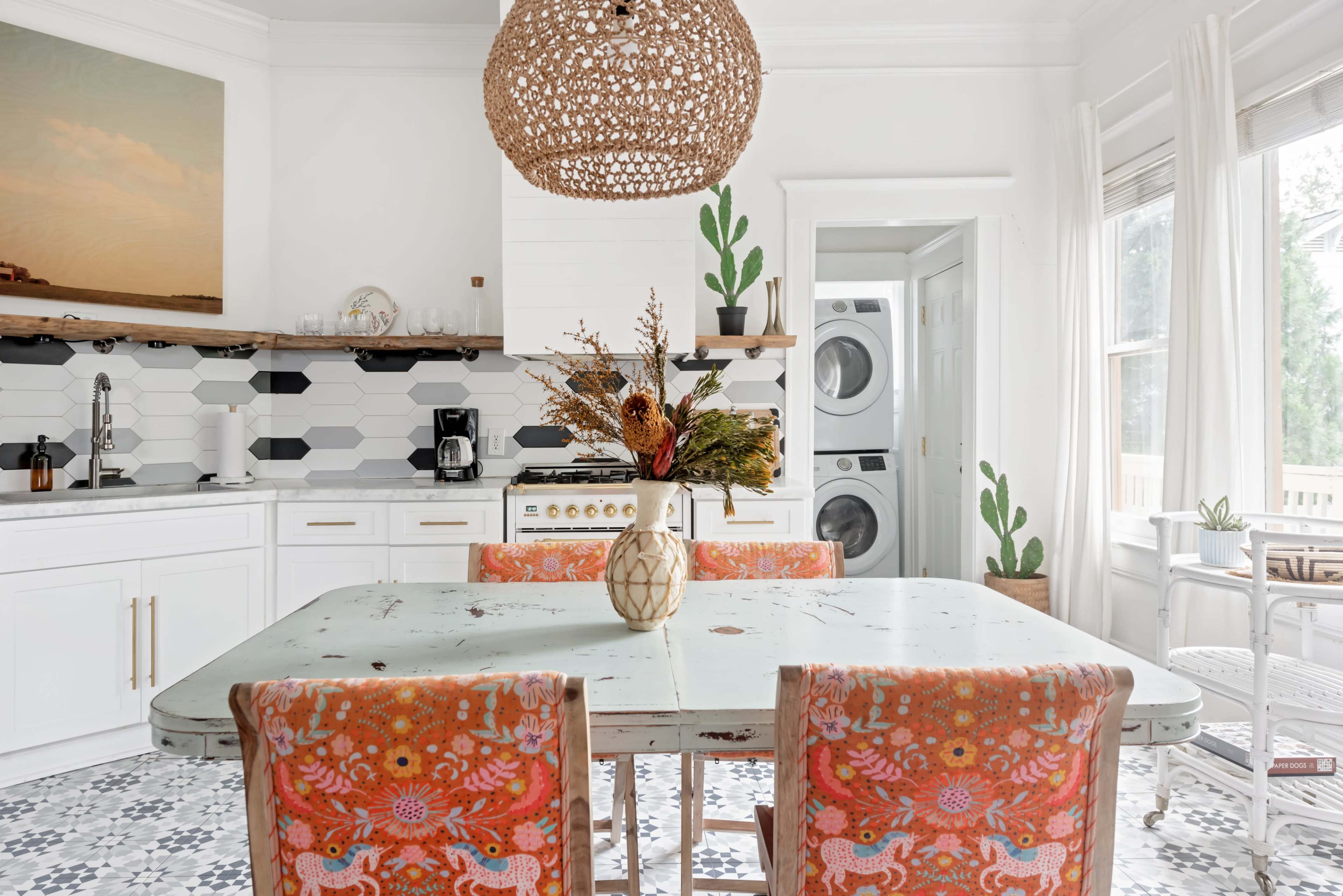 A bright kitchen features a large white table with orange-patterned chairs, a woven pendant light above, and a laundry area visible in the background.