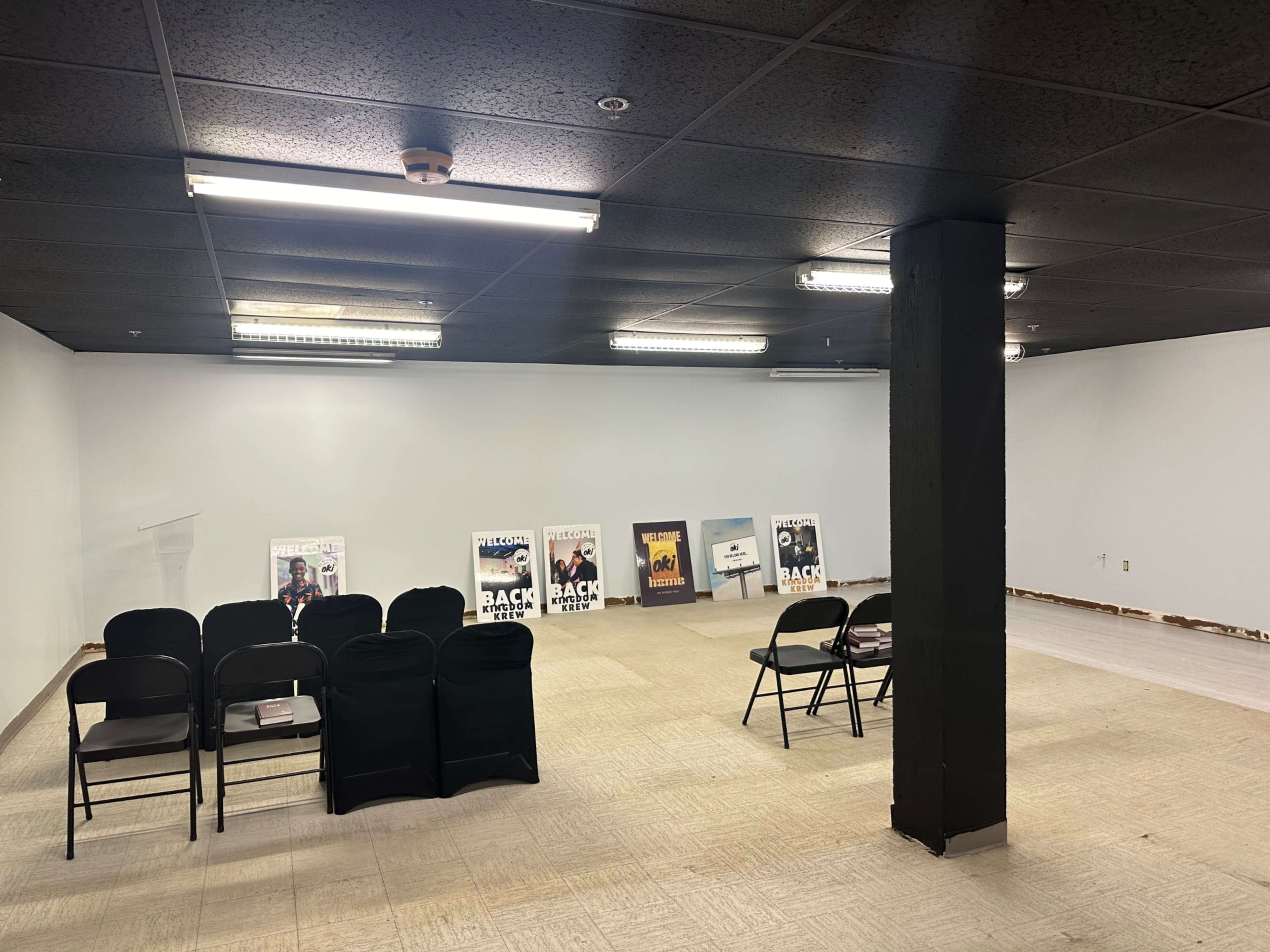 The image shows a large, empty room with a few rows of black chairs facing a blank wall, and several posters displaying various titles leaning against the wall.