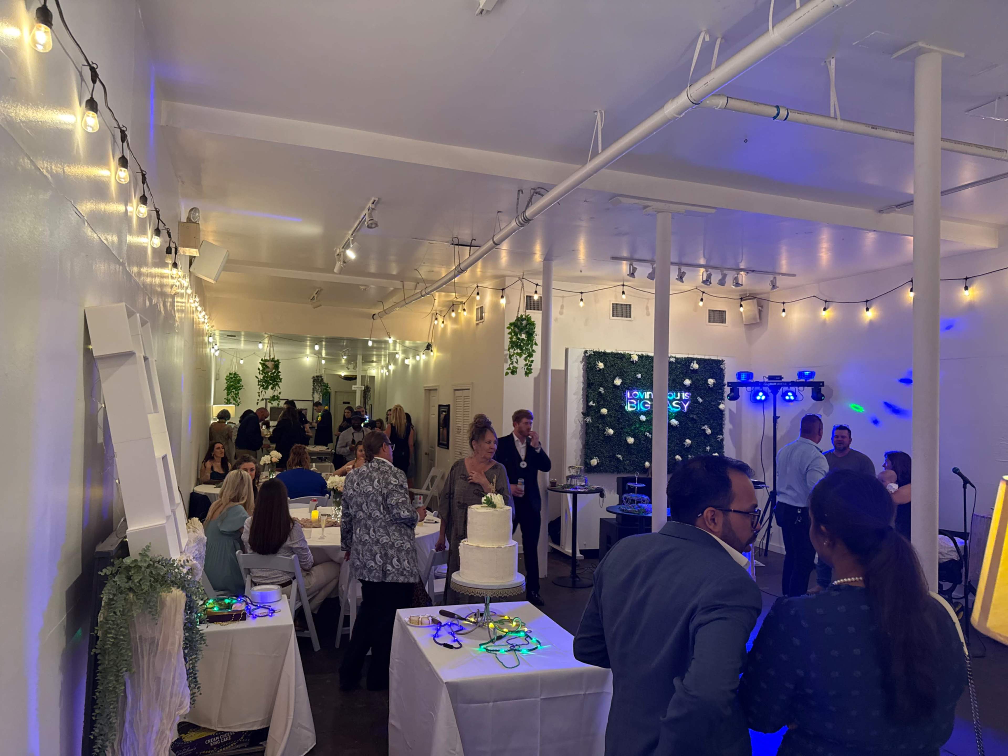 The image shows a lively indoor event space with guests mingling, tables set up, and a cake displayed prominently, accompanied by decorative lighting and a stage area.