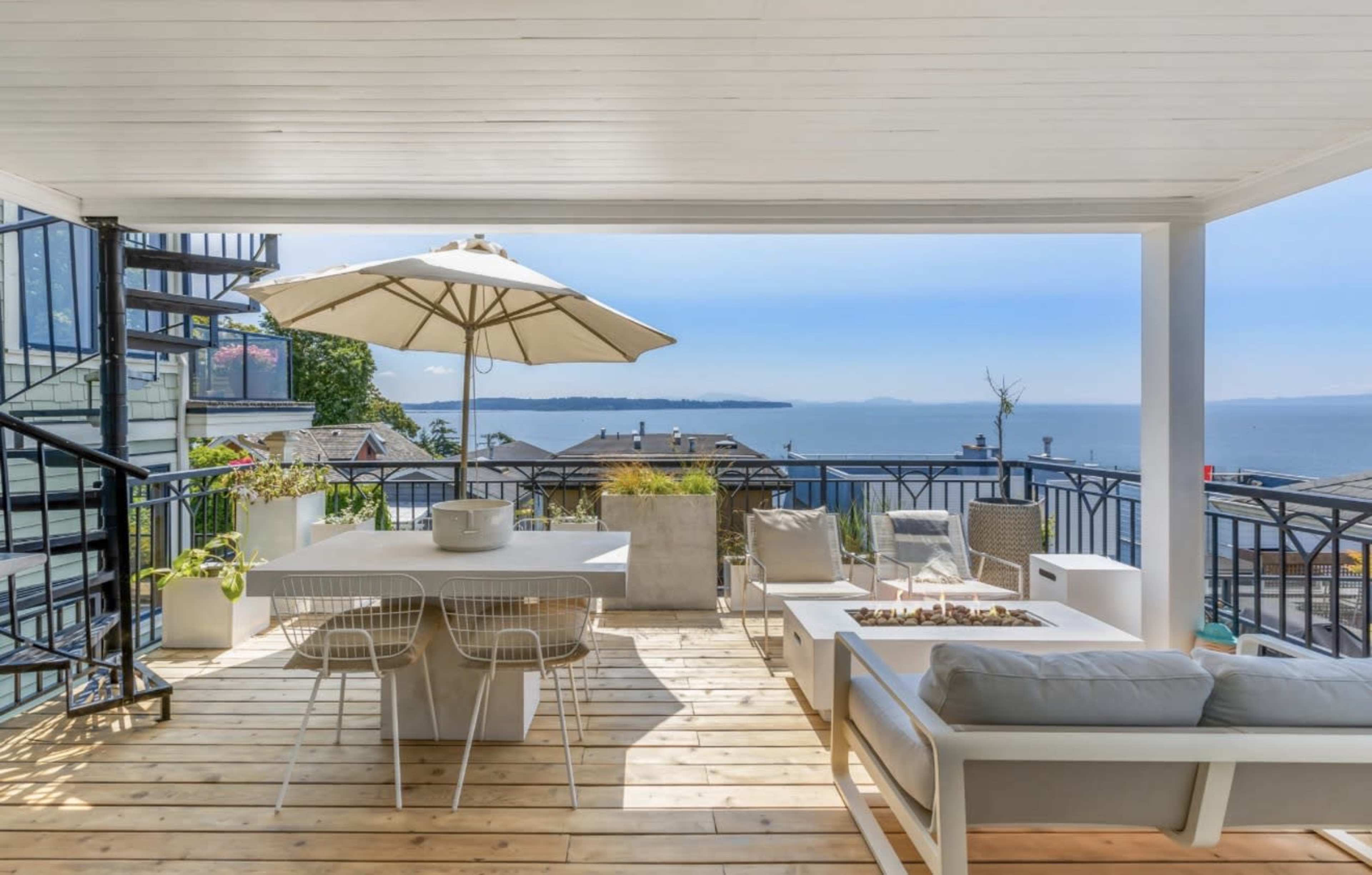 The image depicts a modern outdoor patio with a dining table, lounge seating, and an umbrella, overlooking a body of water and distant land.