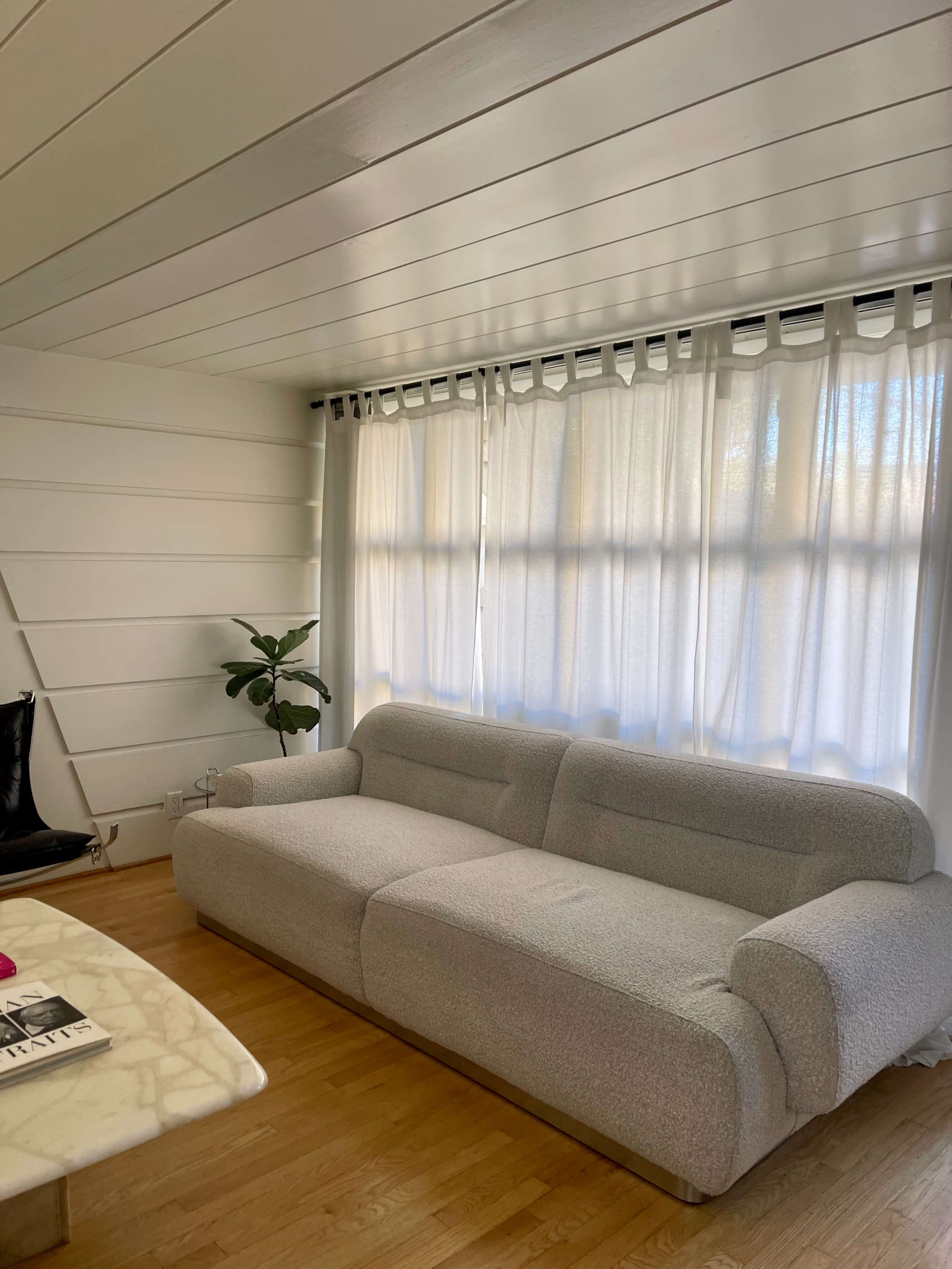 A spacious living room features a gray sofa, a marble coffee table, and large windows covered with white curtains.