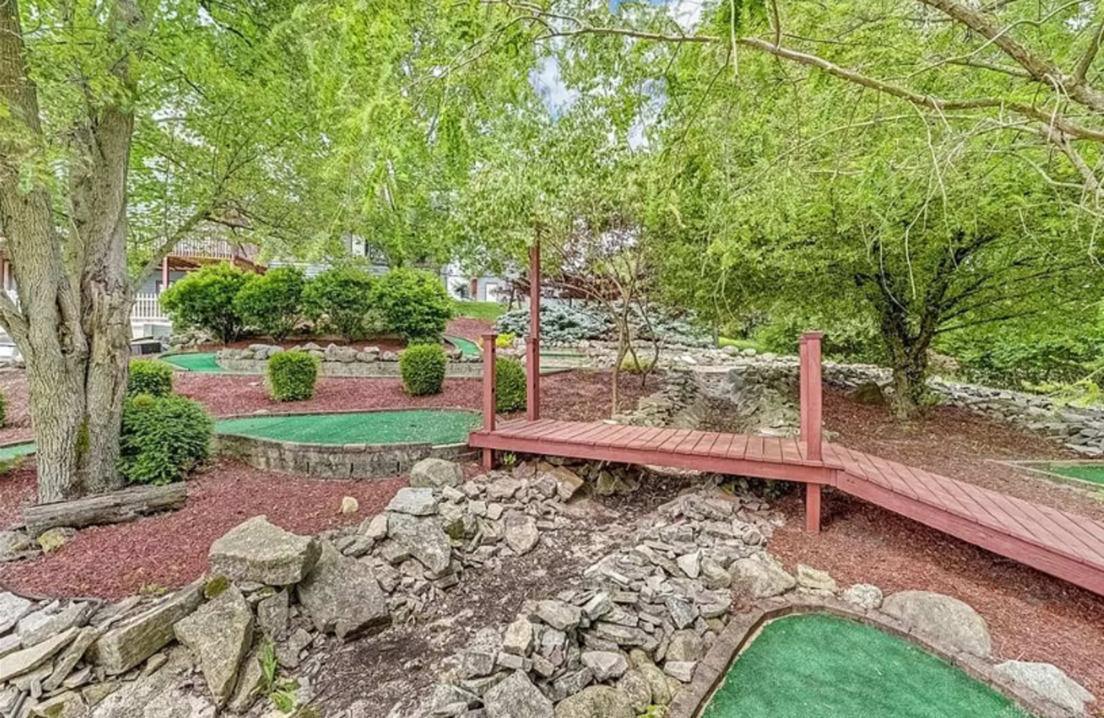 A landscaped backyard with a wooden bridge crossing over rocks and grassy patches surrounded by trees and shrubbery.