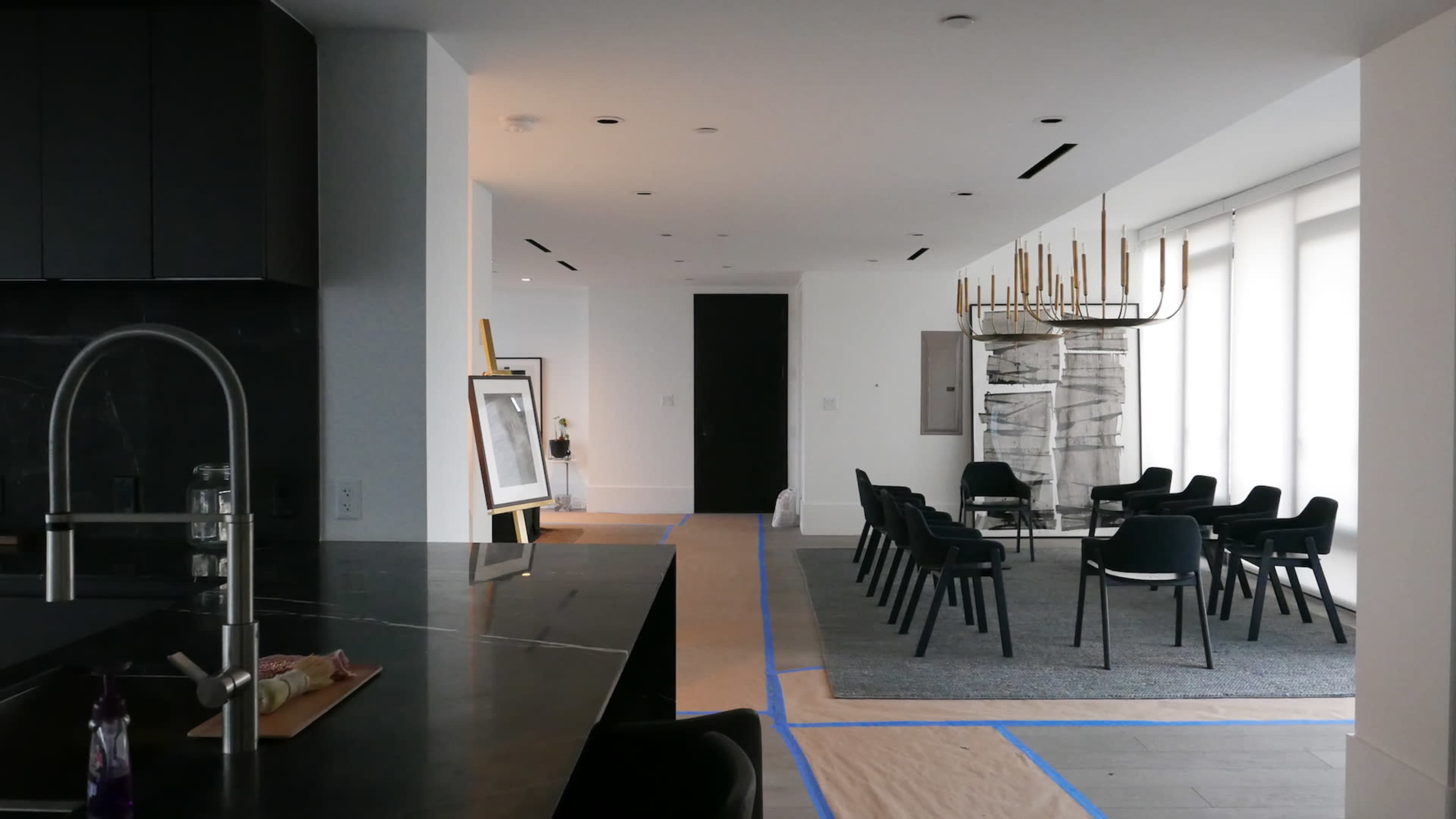 The image shows a modern, partially renovated interior space with a kitchen area to the left and a room featuring a dining setup on the right, and construction materials laid on the floor.