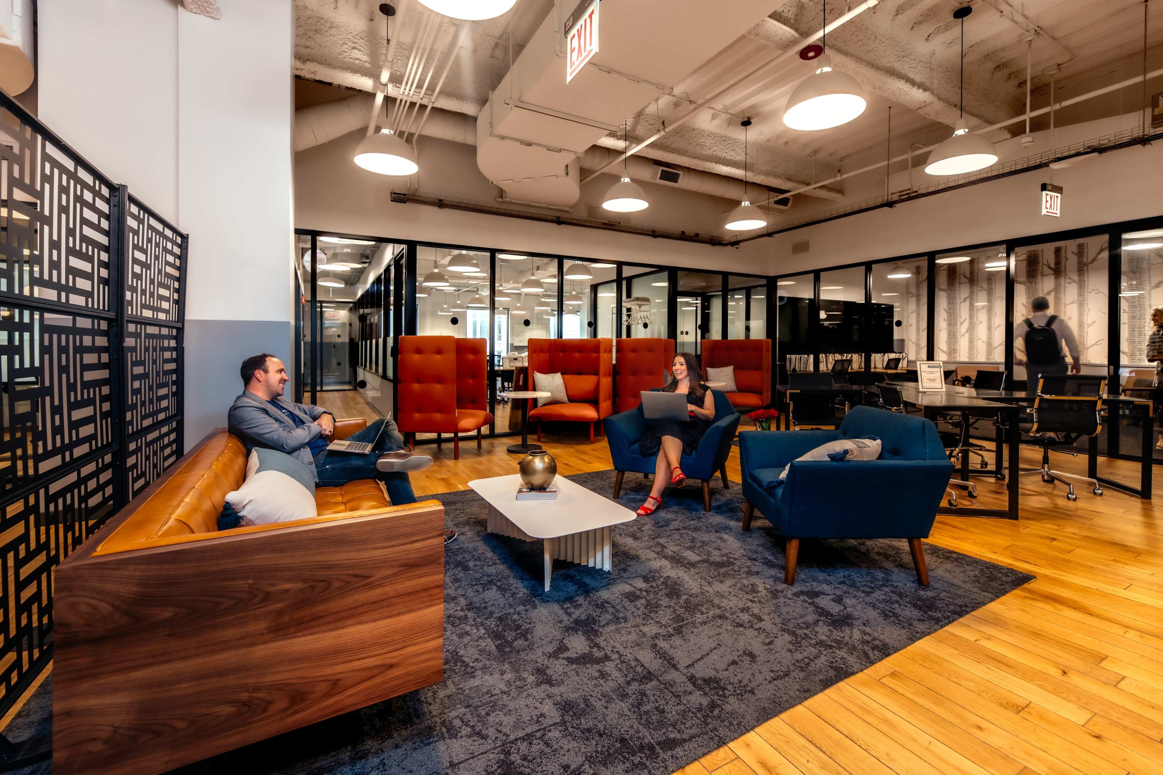 A modern office space features a seating area with orange and blue chairs, a wooden table, and glass-walled meeting rooms in the background.