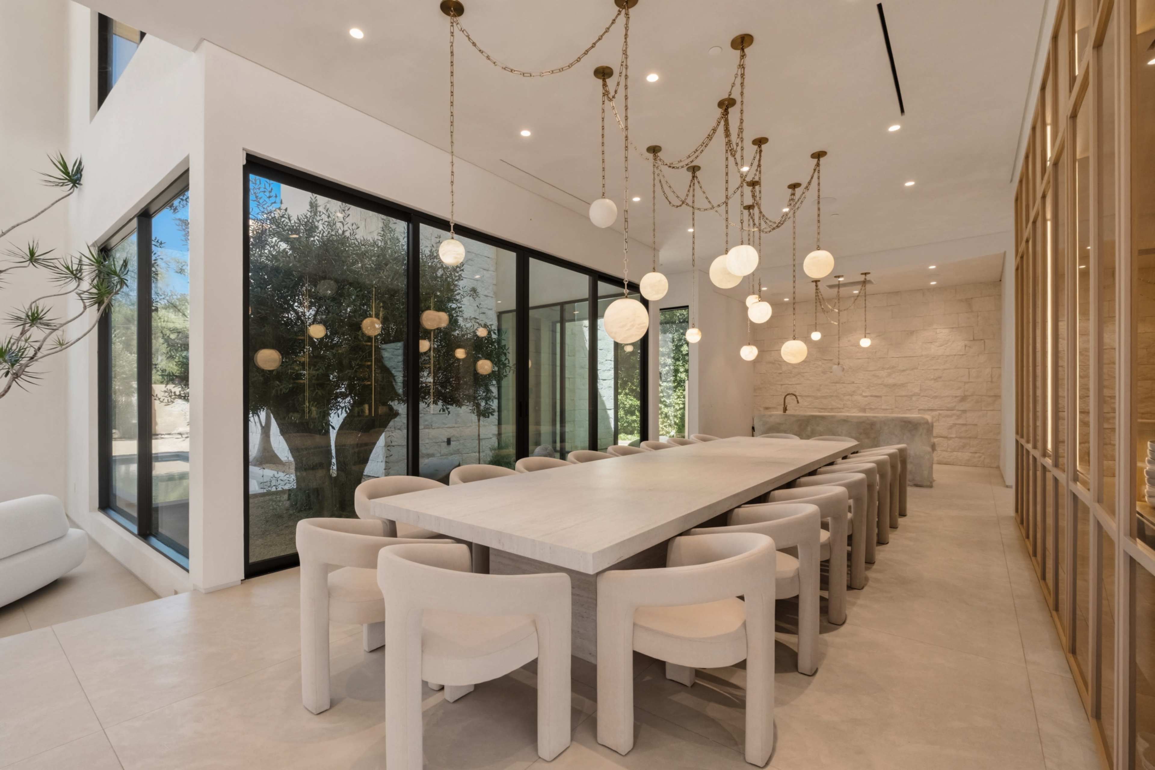 A modern dining area features a large table surrounded by upholstered chairs, illuminated by pendant lights, with large glass windows showcasing an outdoor olive tree.