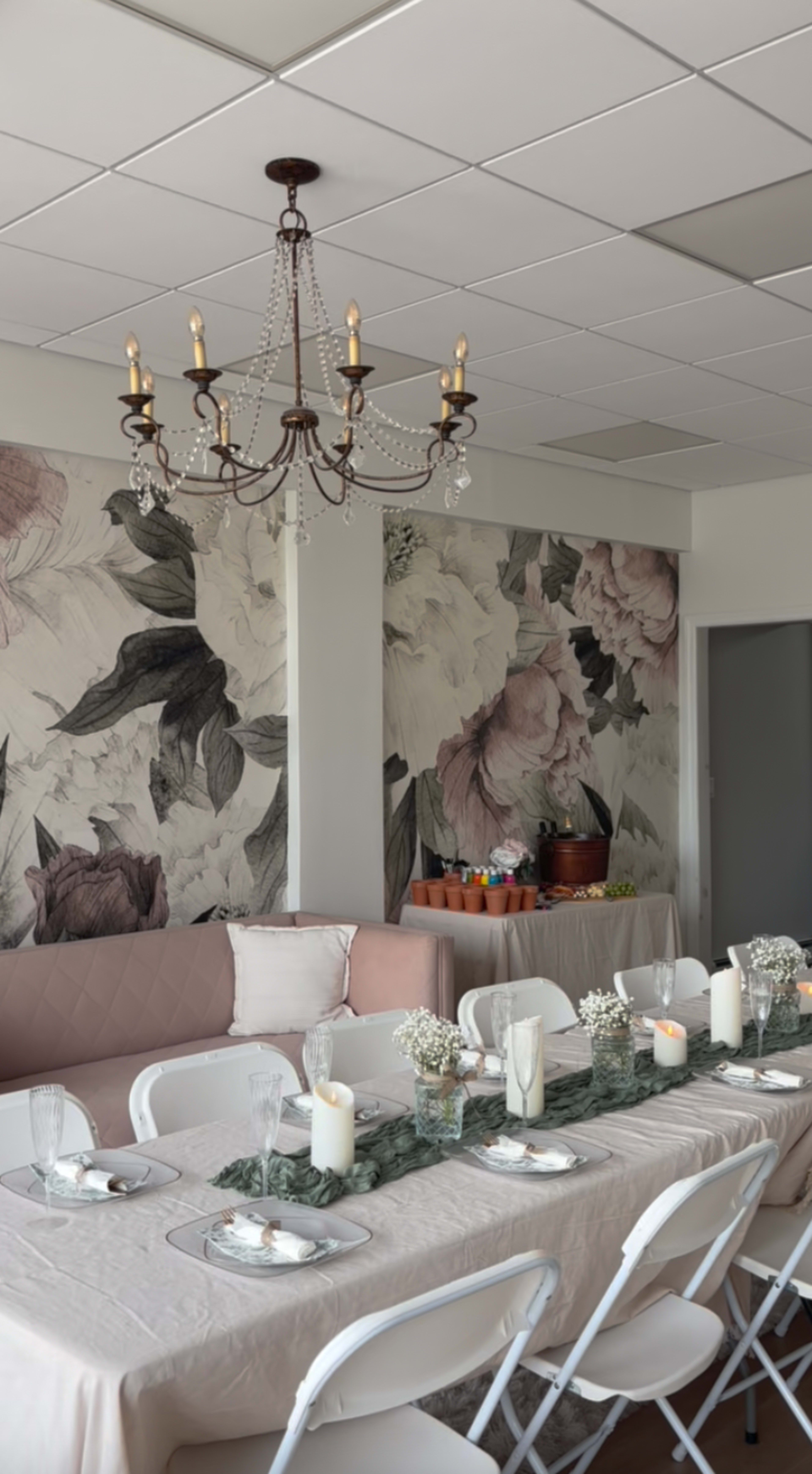 A long dining table is set with candles and tableware in a room featuring floral wallpaper and a chandelier overhead.