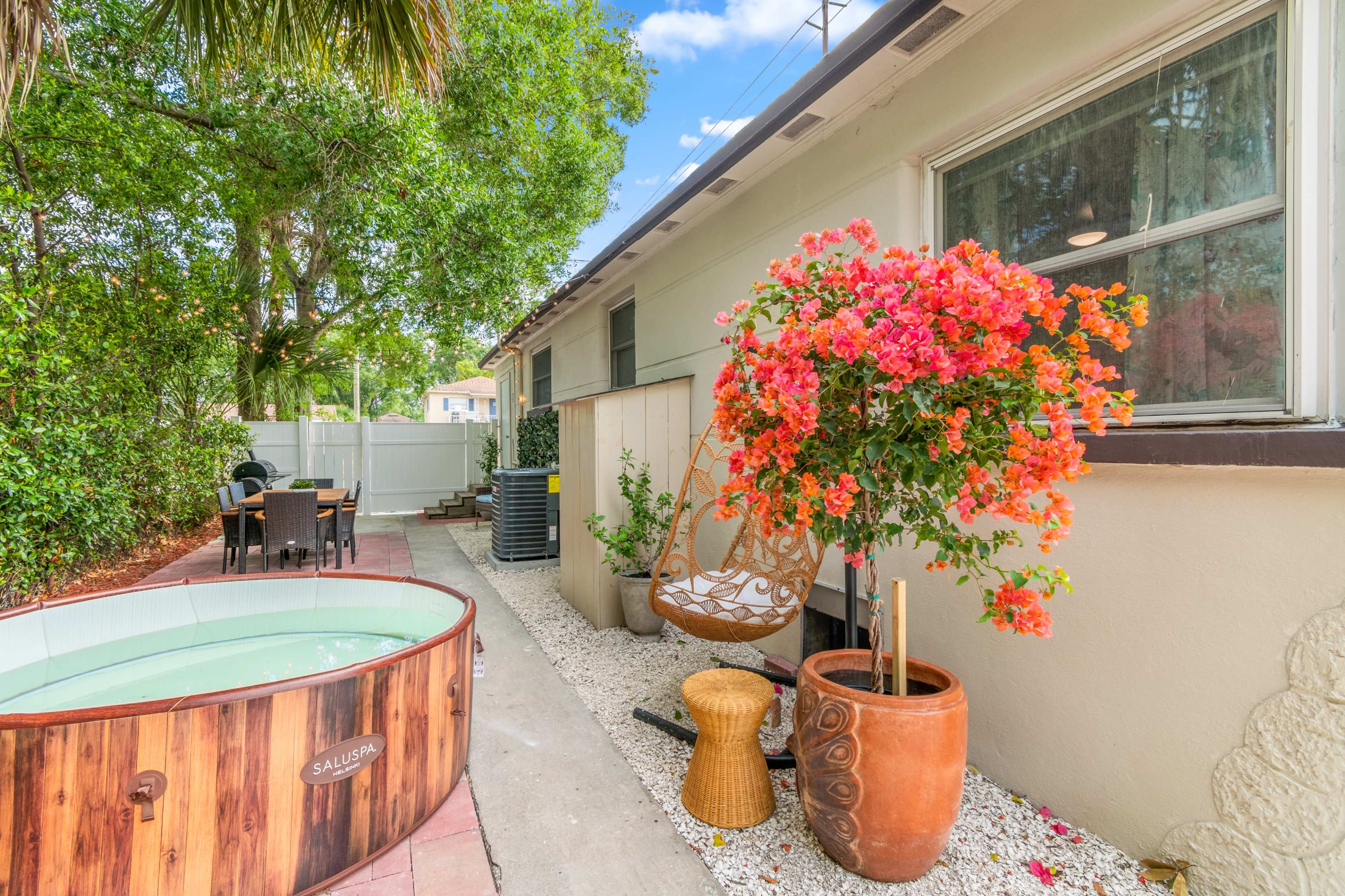 A backyard scene with a wooden hot tub, colorful flowering plants, and outdoor seating near a house.